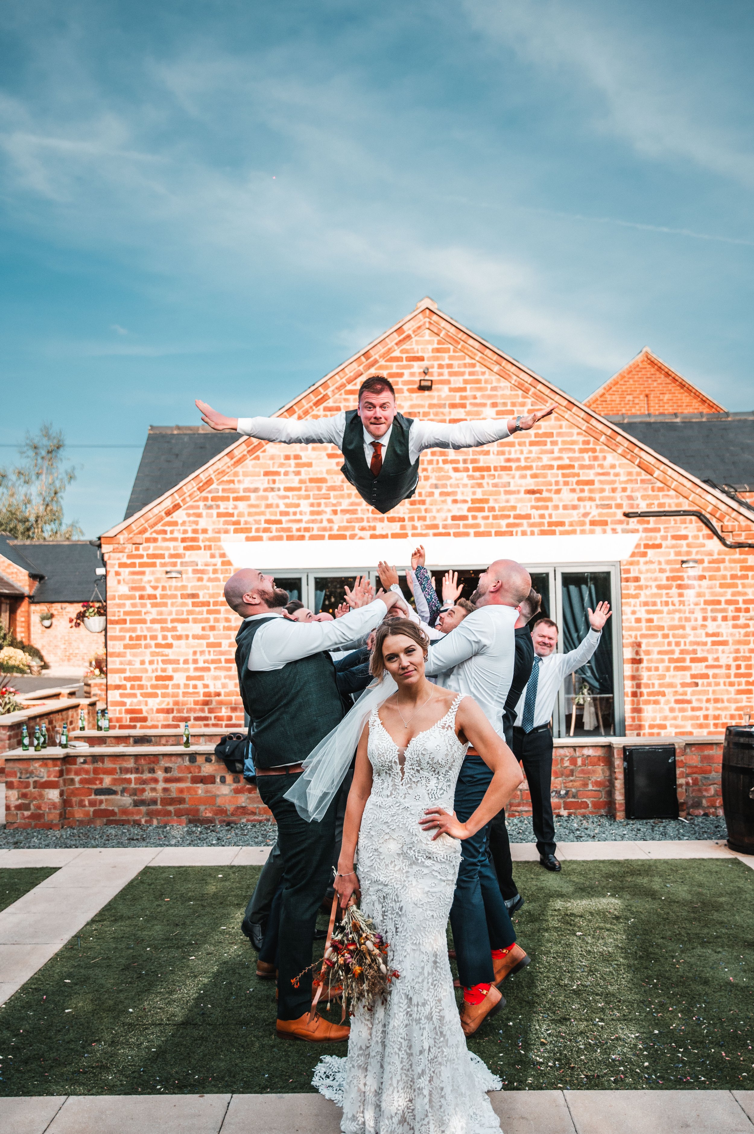 A bride standing in front of a group of people celebrating at a wedding reception outdoors during daytime, with a man jumping over the crowd with arms outstretched and others in the background cheering and smiling.