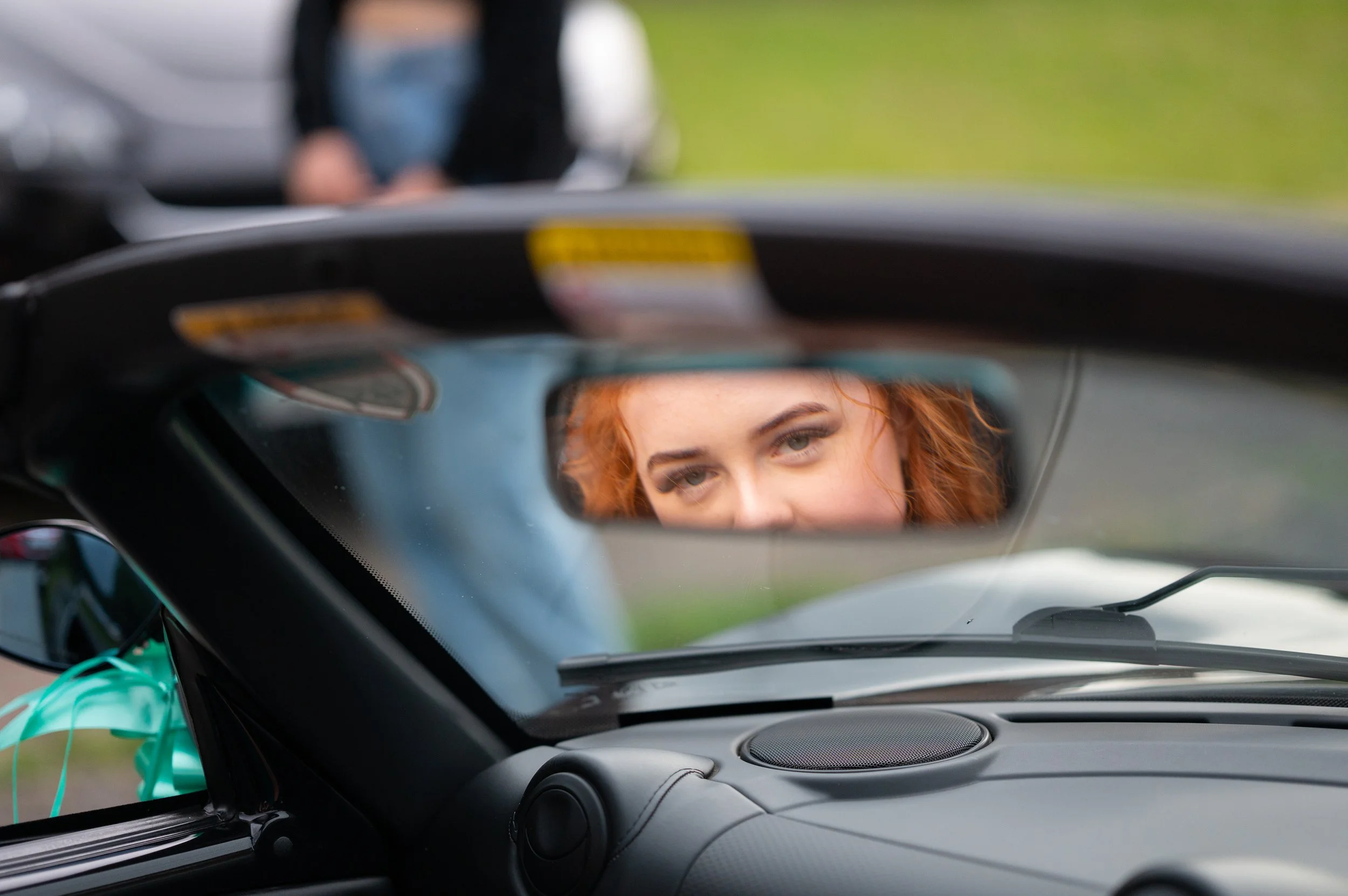 Close-up of a woman's face reflected in a car's rearview mirror, with her red hair and green eyes visible. The image is taken from inside a vehicle, focusing on the mirror.