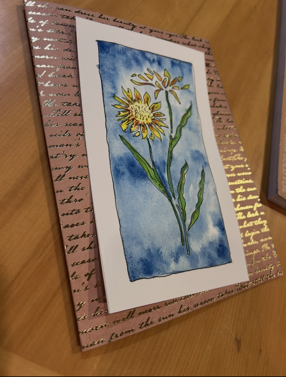 A handmade card with a watercolor illustration of two yellow flowers, set against a blue sky background.