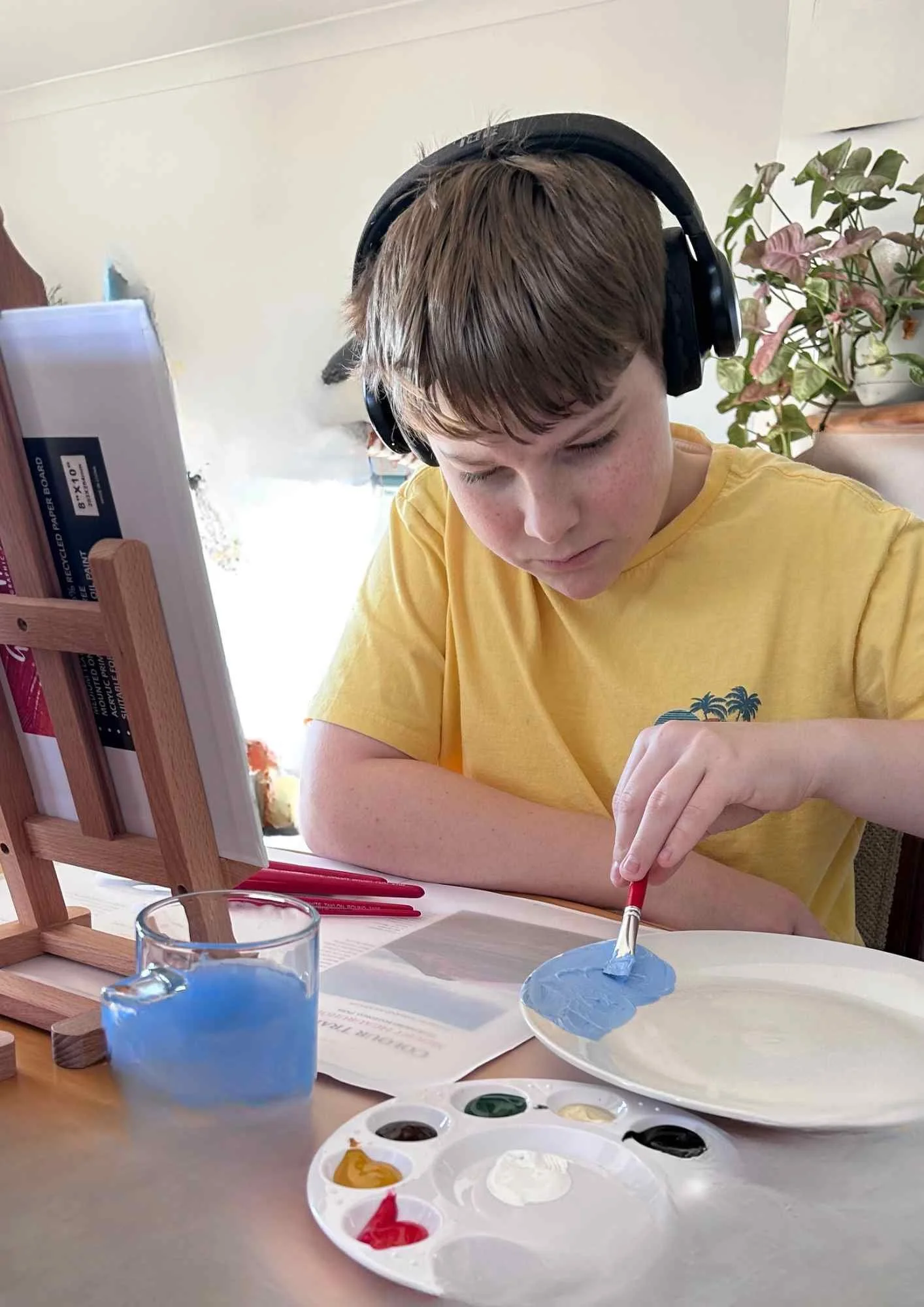 A boy painting with blue paint on a canvas, wearing a yellow shirt and black headphones, sitting at a table with painting supplies, including a palette with paint and a glass of blue water. There's a plant in the background.