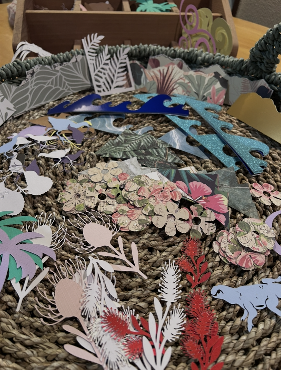 Colorful paper cutout decorations for crafting, including various floral, animal, and nature-themed shapes, arranged on a woven surface with a wooden storage box in the background.