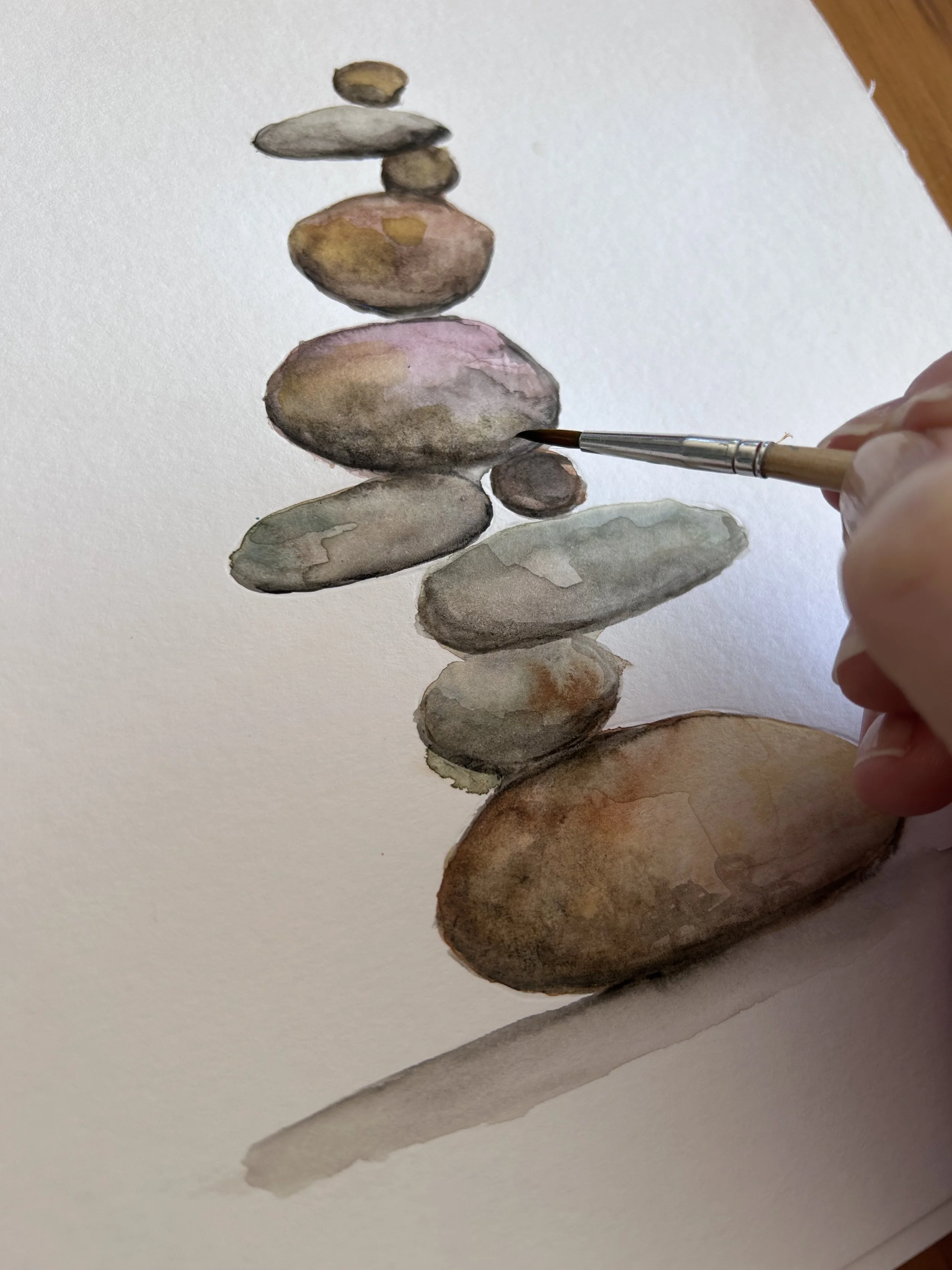 Watercolor painting of a stack of smooth, multicolored stones on white paper, with a hand holding a paintbrush adding details.
