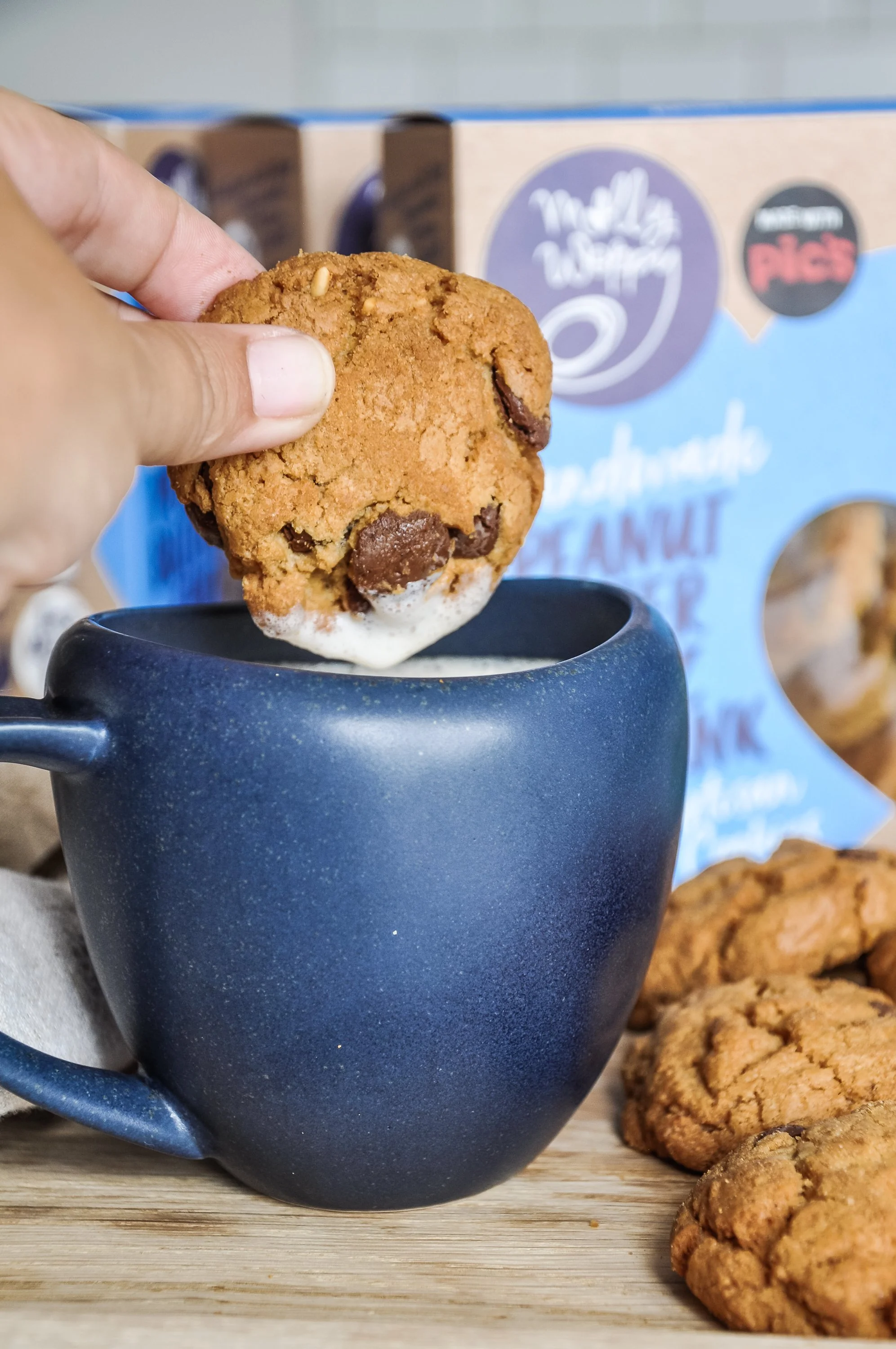 A person holding a chocolate chip cookie over a dark blue mug, with more cookies and a box of cookies in the background.