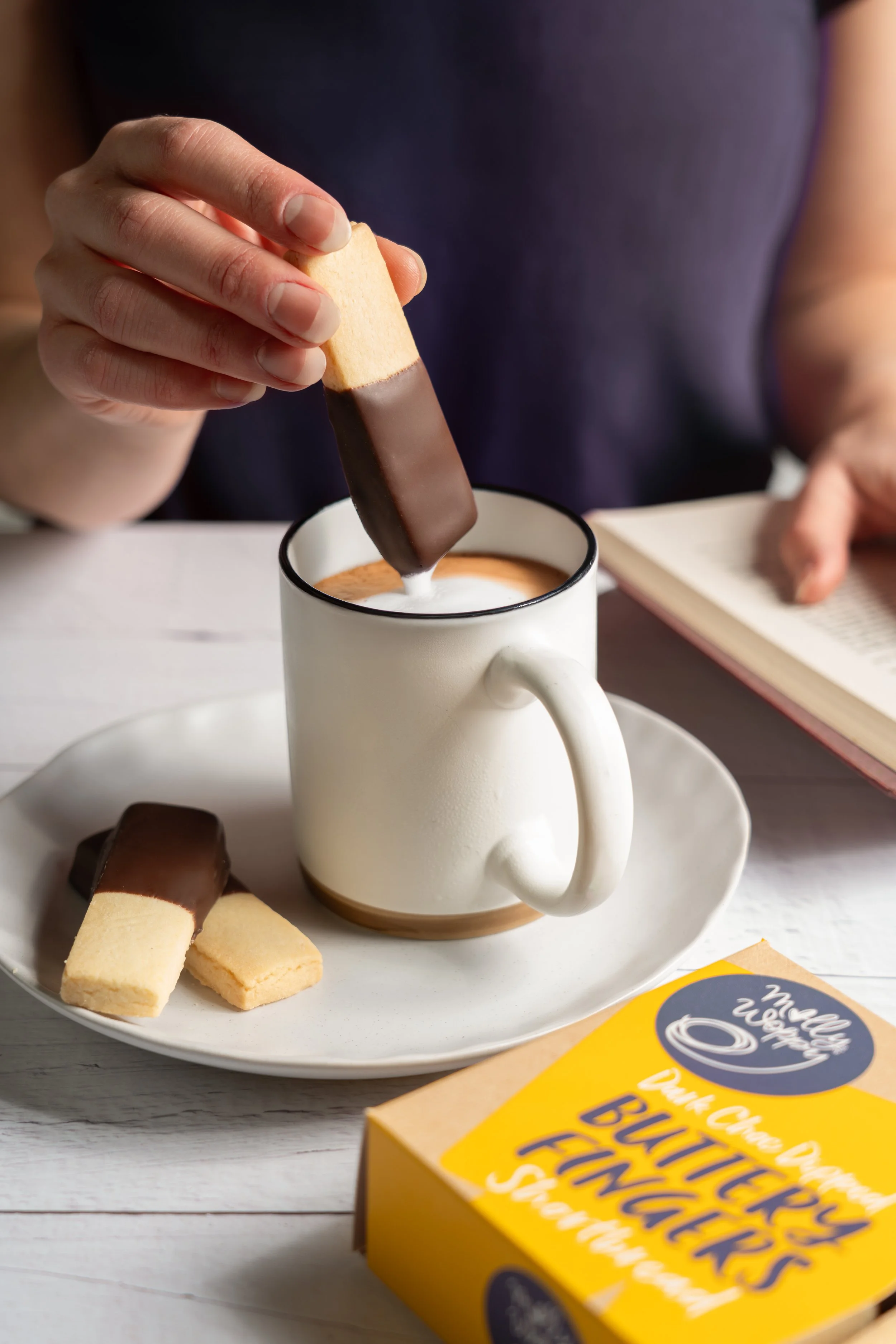 Person dipping a cookie in a mug of milk with cookies on a white plate in front, a box of cookies on a white table.
