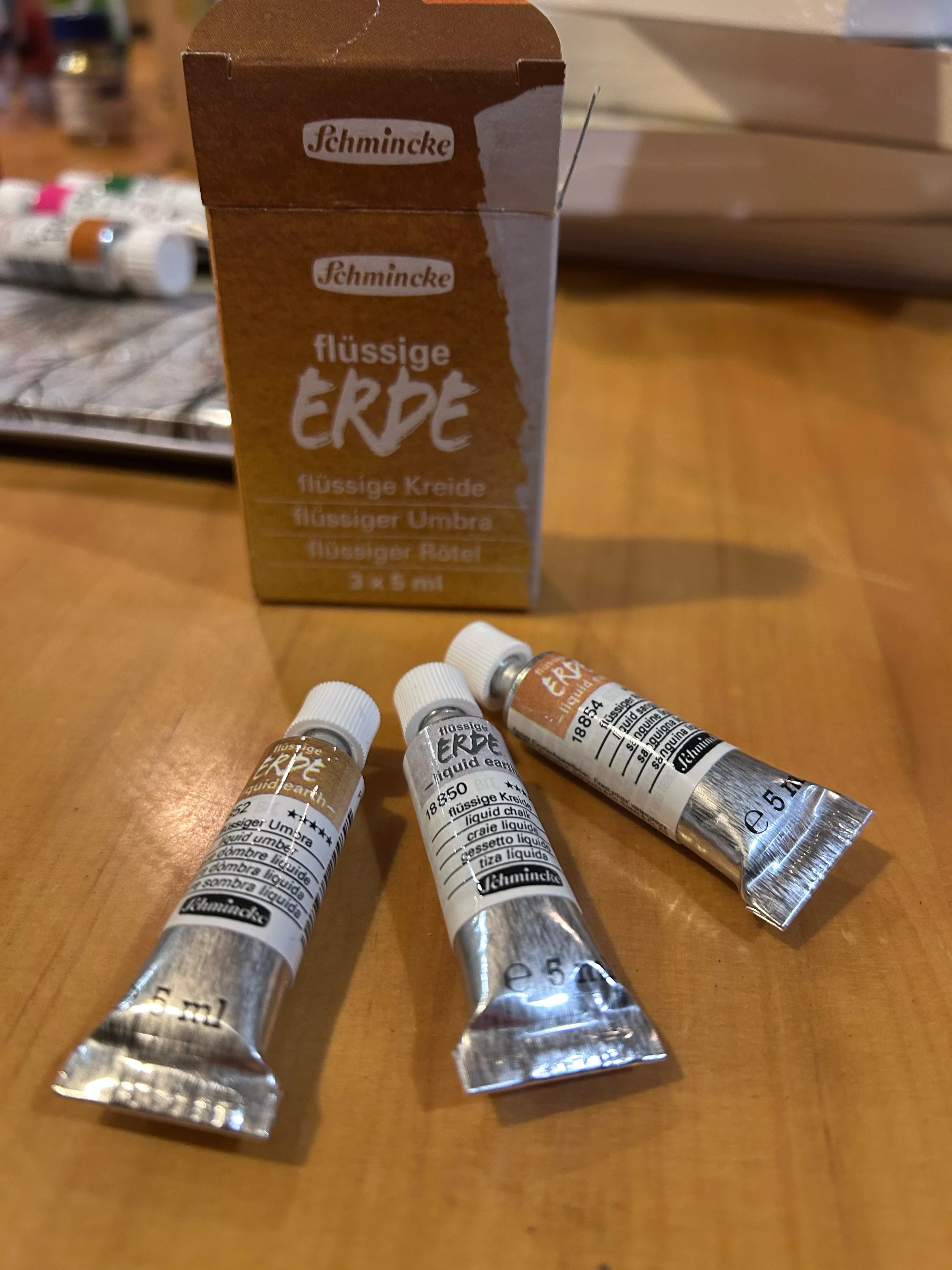 Three tubes of Schmincke liquid earth paint with a box behind them on a wooden surface.