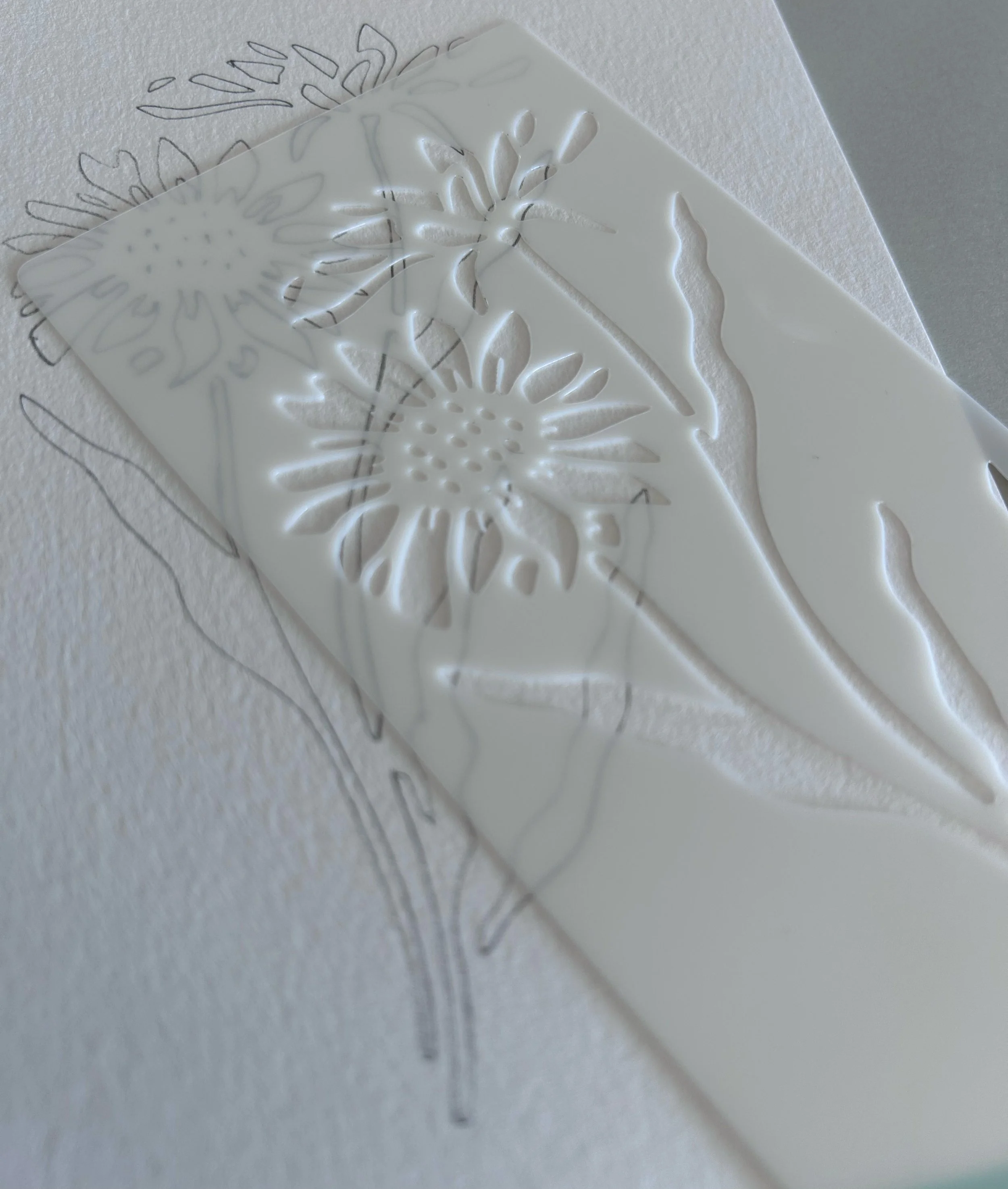 A clear acrylic stencil with floral cutouts resting on a piece of paper with a floral outline drawing, both placed on a white textured surface.