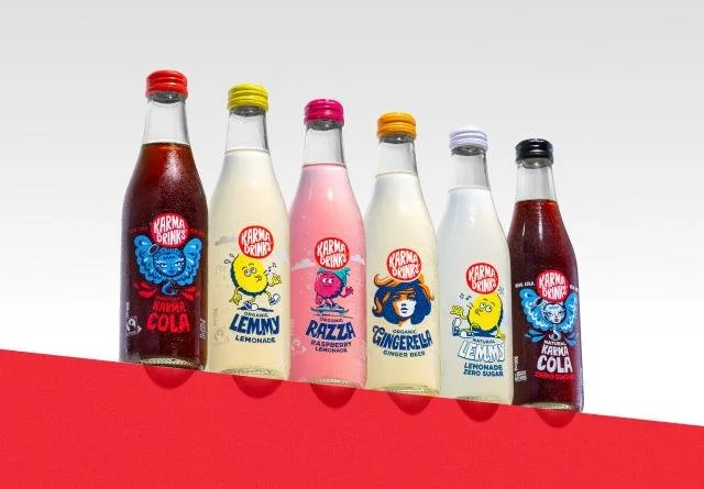 Six bottles of Karma Drinks soda arranged in a row on a red surface, featuring flavors such as colas, lemony, raspberry, gingerbar, and lemon zero sugar.