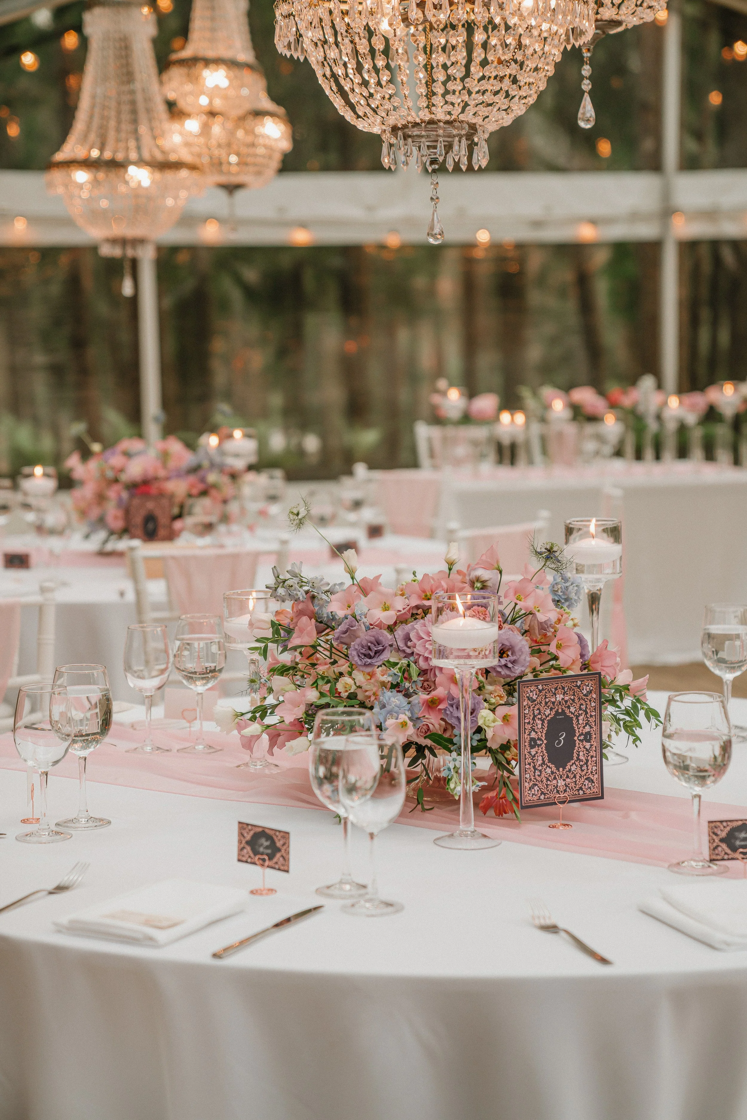 Elegant outdoor wedding reception table decorated with a large floral centerpiece, candles, and glassware, under hanging chandeliers with a forest background.