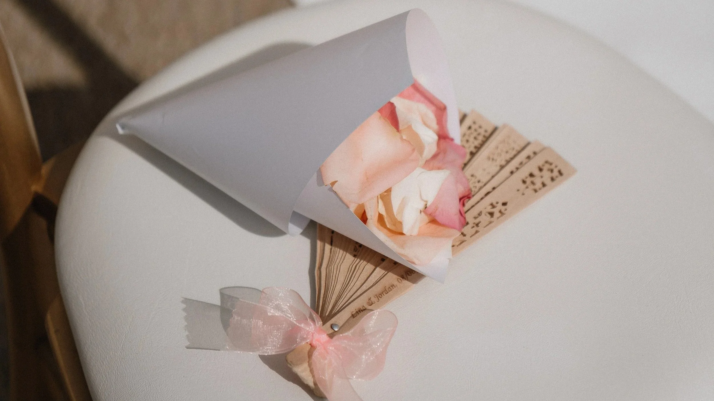 A small bouquet wrapped in white paper with pink and white rose petals, tied with a pink ribbon, resting on a wooden display with engraved text on a white round surface.