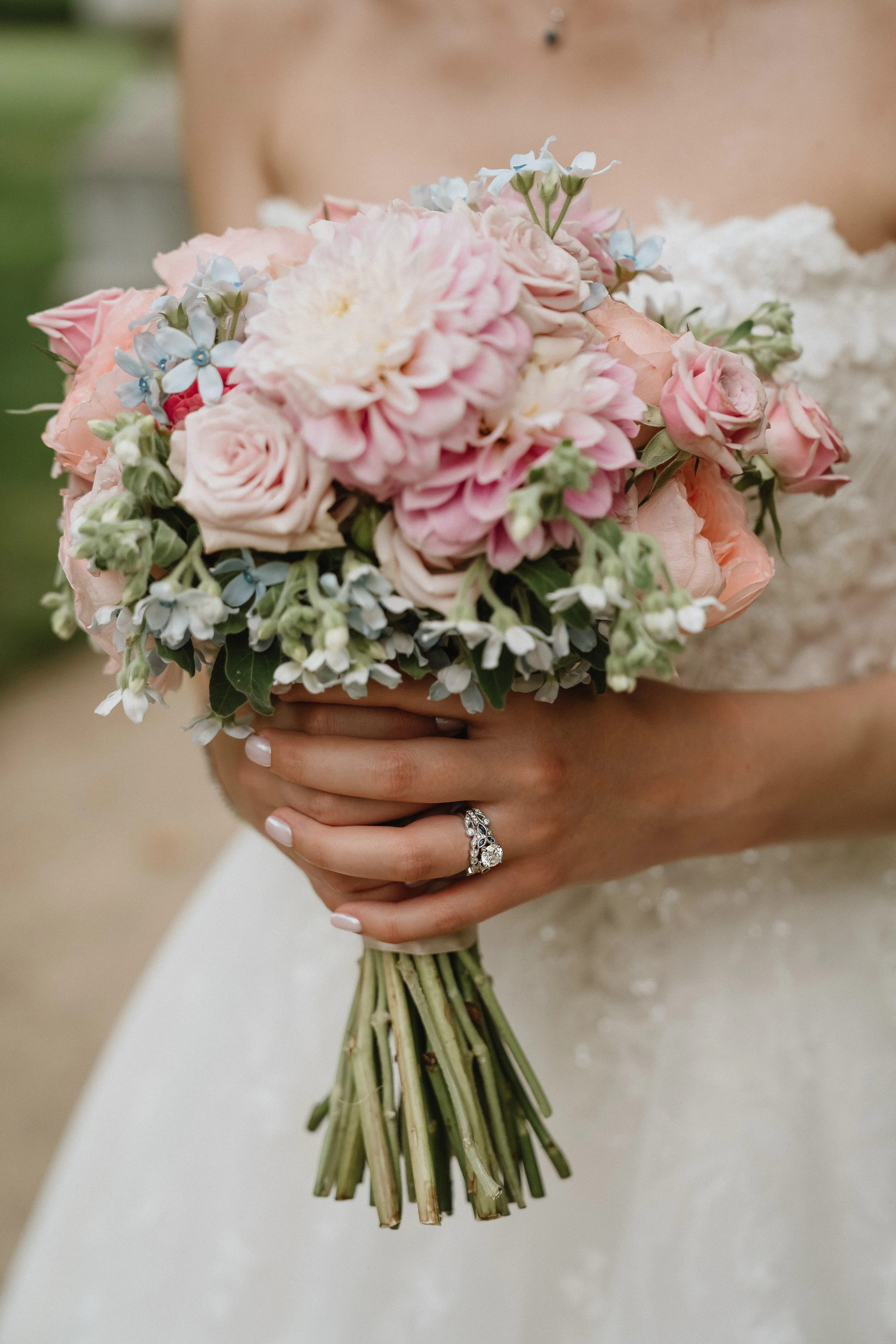 A bride holding a bouquet of pink, white, and blue flowers with a diamond ring on her finger.