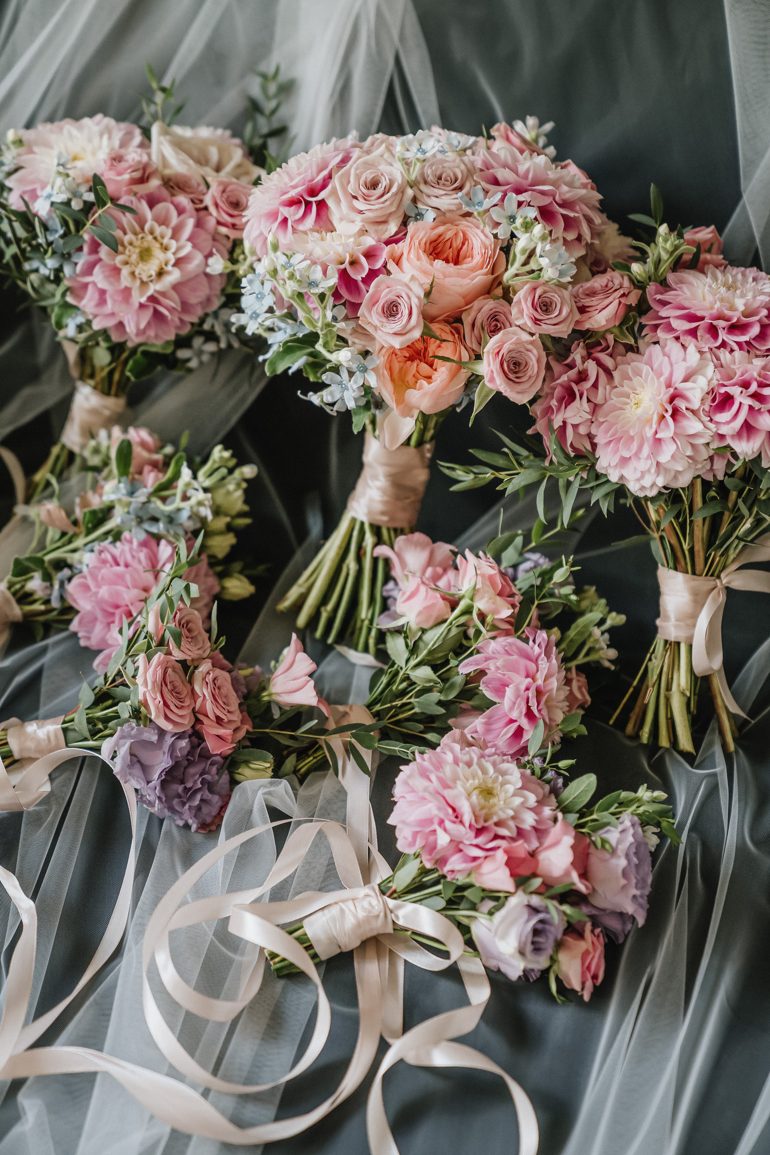 Multiple bouquets of pink and peach roses, dahlias, and other flowers wrapped with satin ribbons, arranged on a sheer gray fabric backdrop.