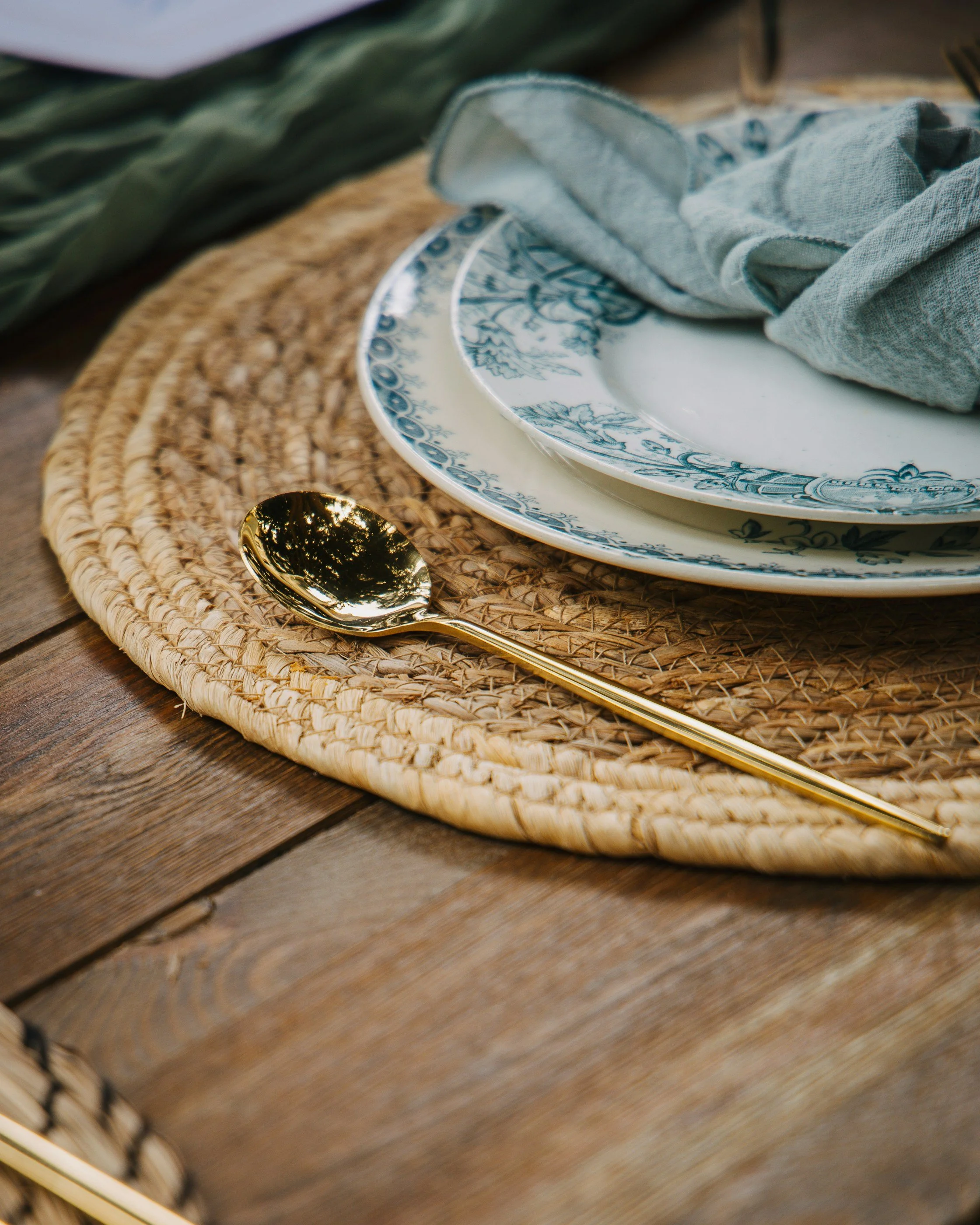 Close-up of a table setting with a woven placemat, blue and white patterned plates, a gray cloth napkin, and a shiny gold spoon, on a wooden surface.