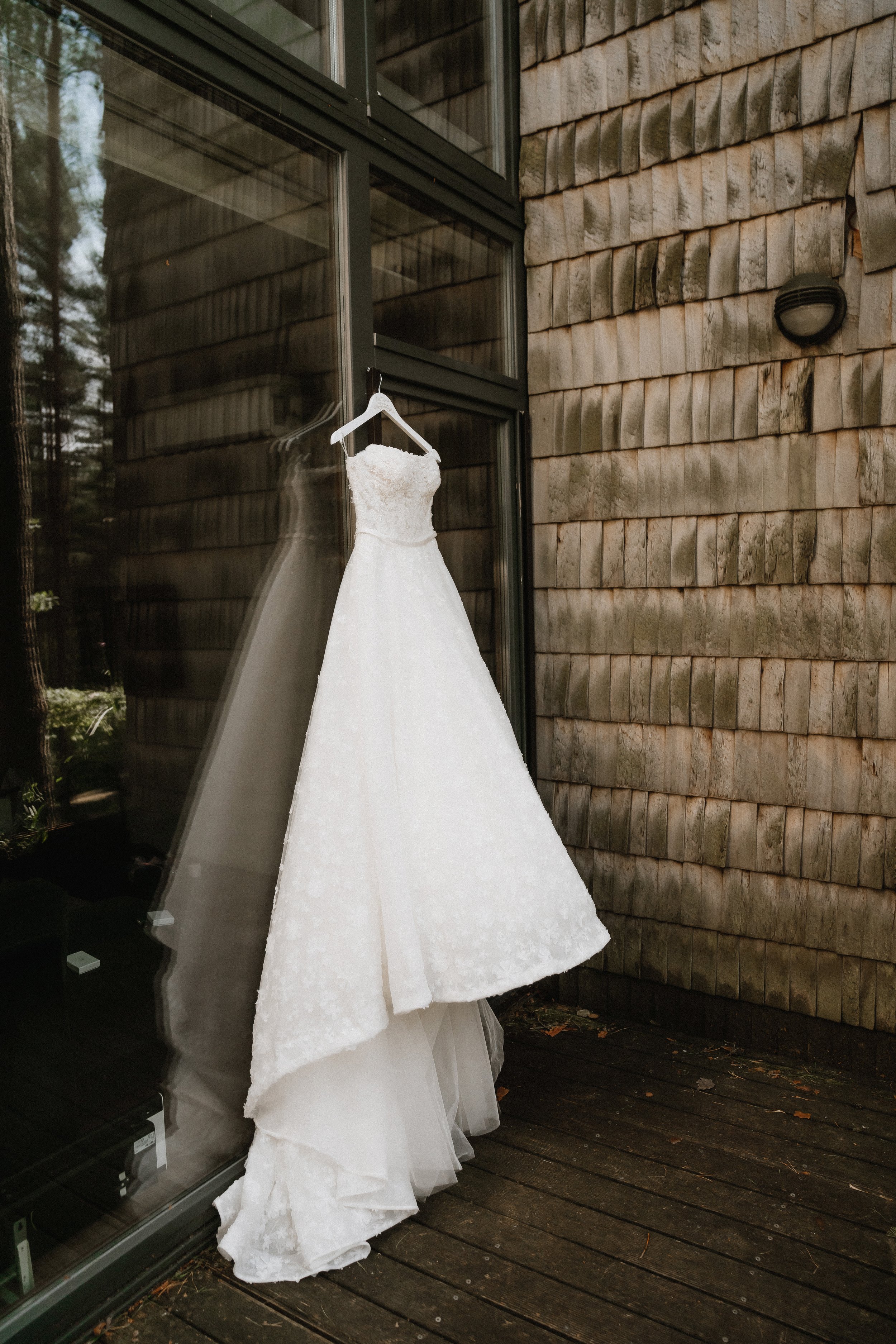 A white wedding dress hanging on a hanger outside a building with wooden siding and large glass windows. The dress is reflected in the glass.