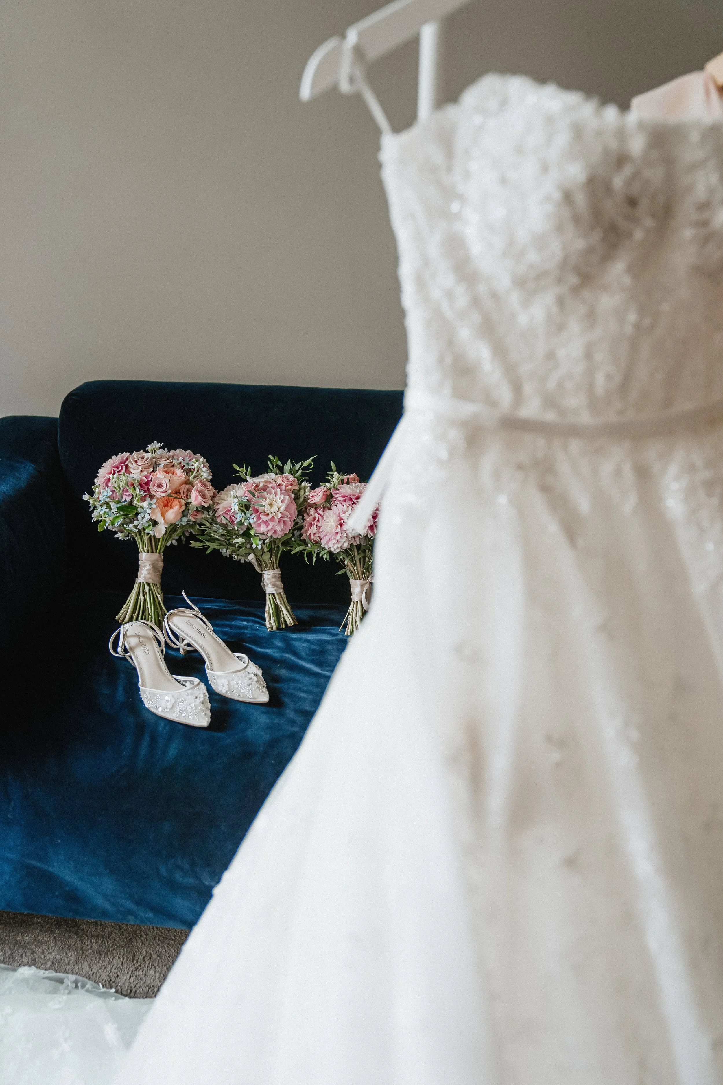 A wedding dress, white with lace details, is partially visible in the foreground. In the background, on a dark blue velvet surface, there are several bouquets of pink and white flowers, and a pair of white lace wedding shoes.