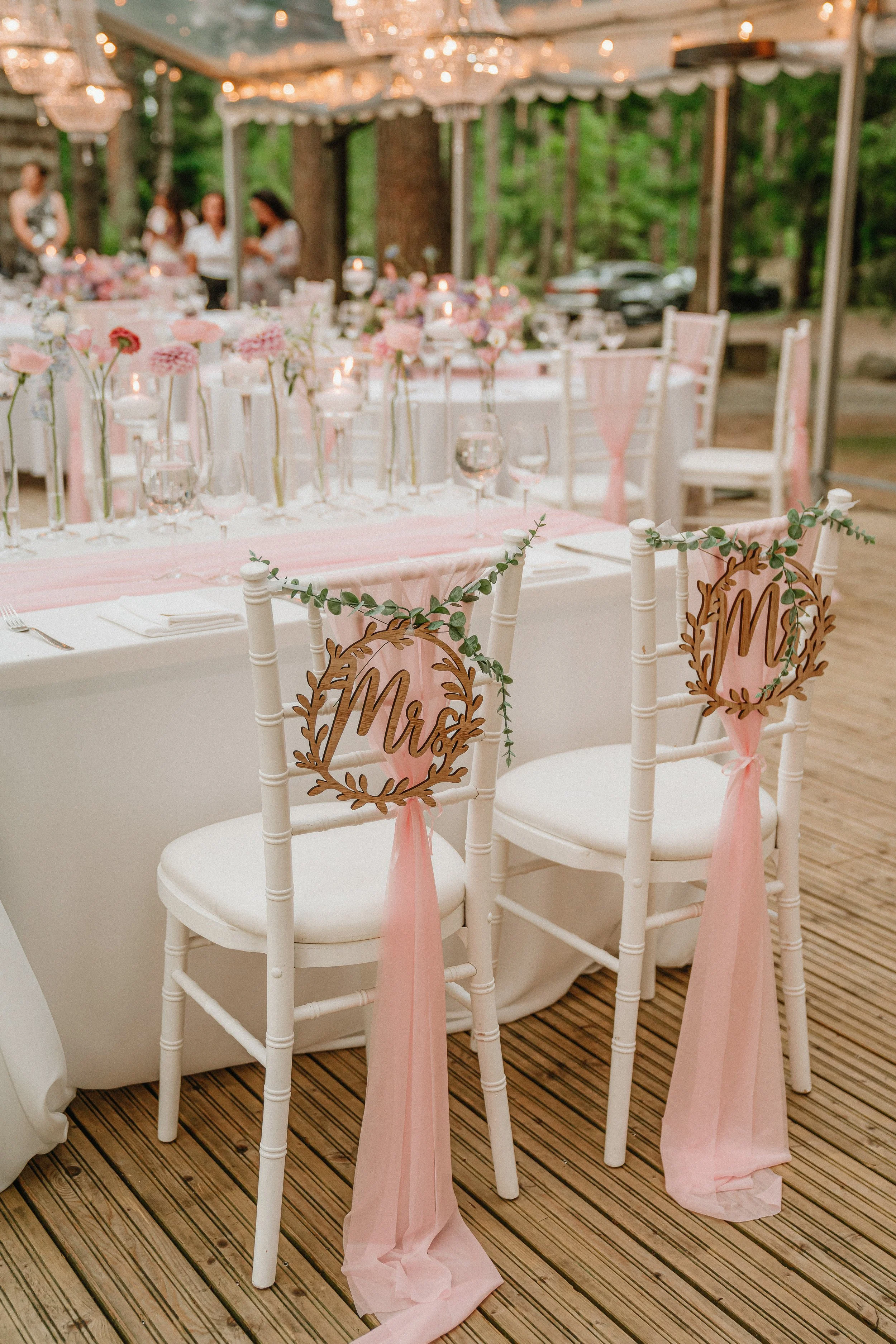Elegant outdoor wedding reception setup with white chairs decorated with pink ribbons and greenery, wooden 'Mr & Mrs' signs, pink flowers, and candle decorations on tables, under string lights in a wooded area.