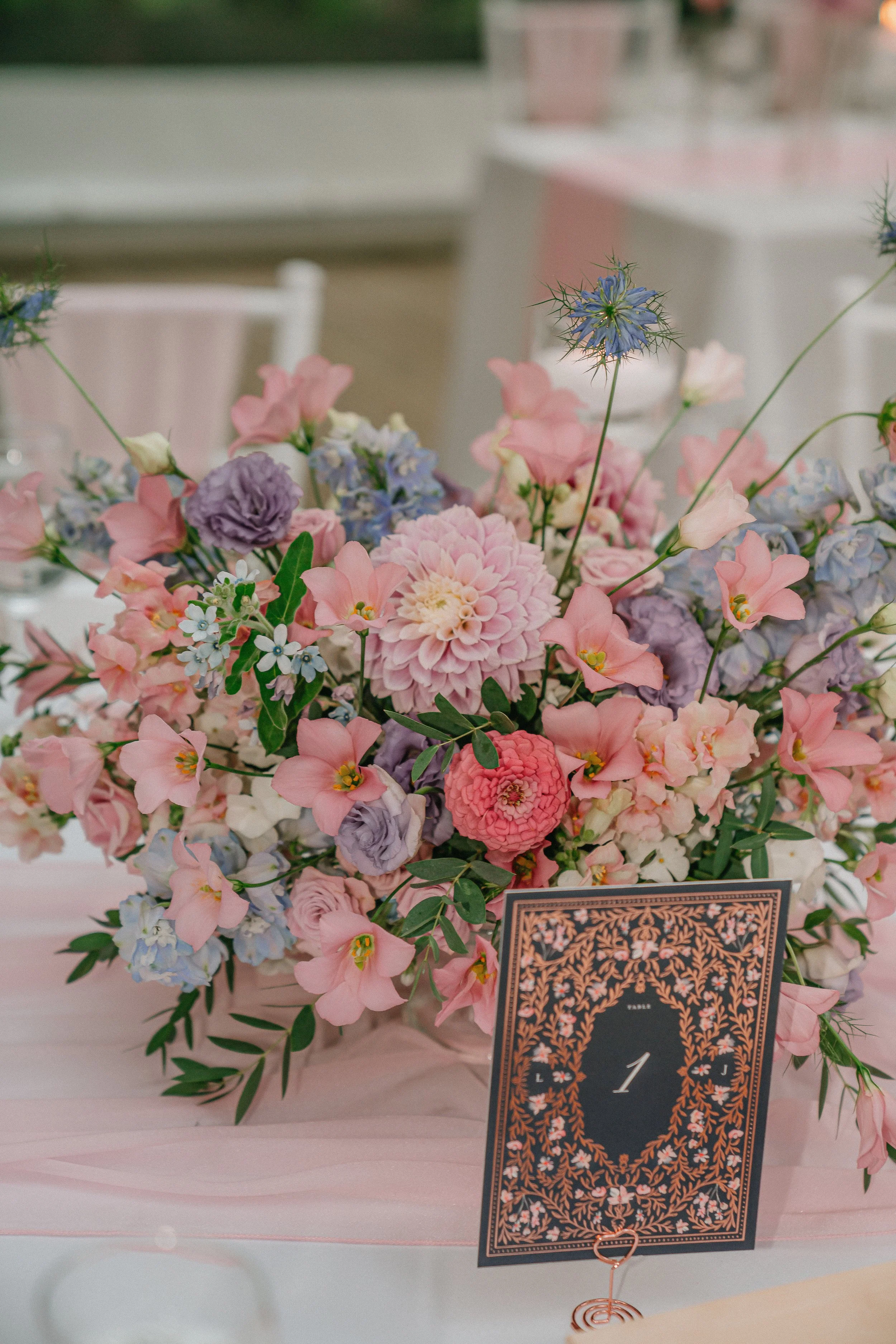 A floral centerpiece with pink, purple, and blue flowers on a table, accompanied by a decorative table number card with intricate gold and floral design, and a small copper wire holder.