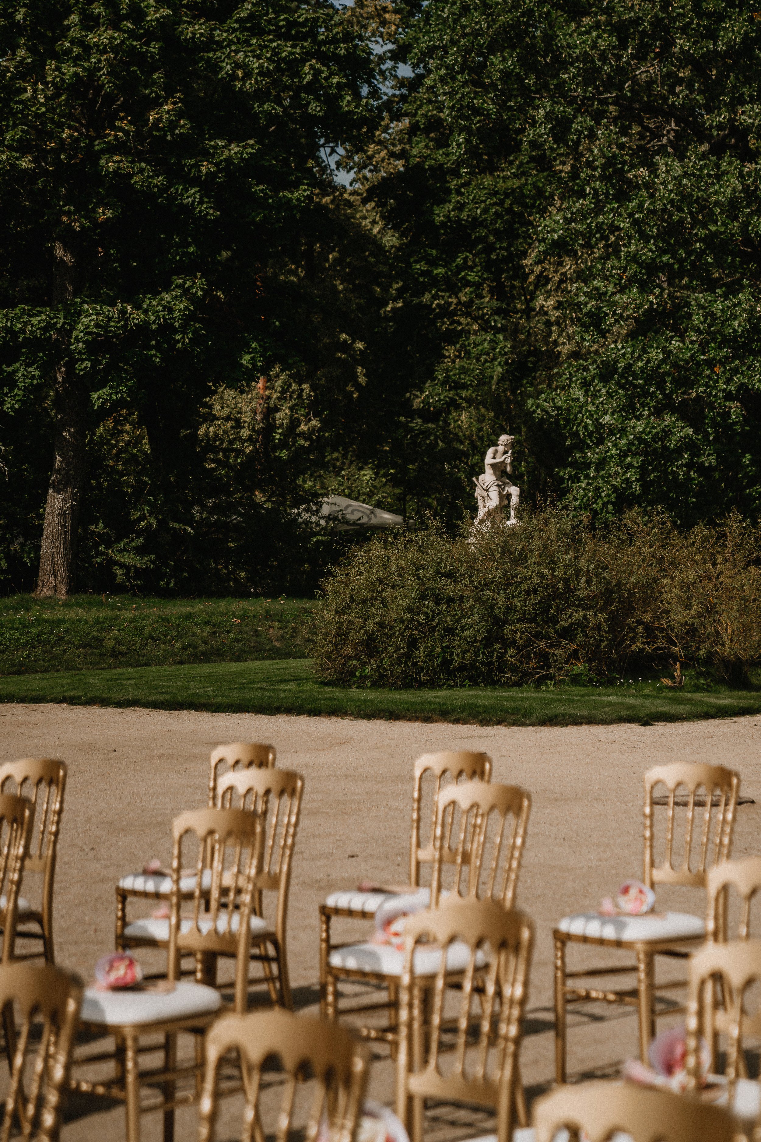 Rows of beige chairs arranged outdoors on a sandy ground, with a backdrop of green trees and a statue of a seated woman in a garden.