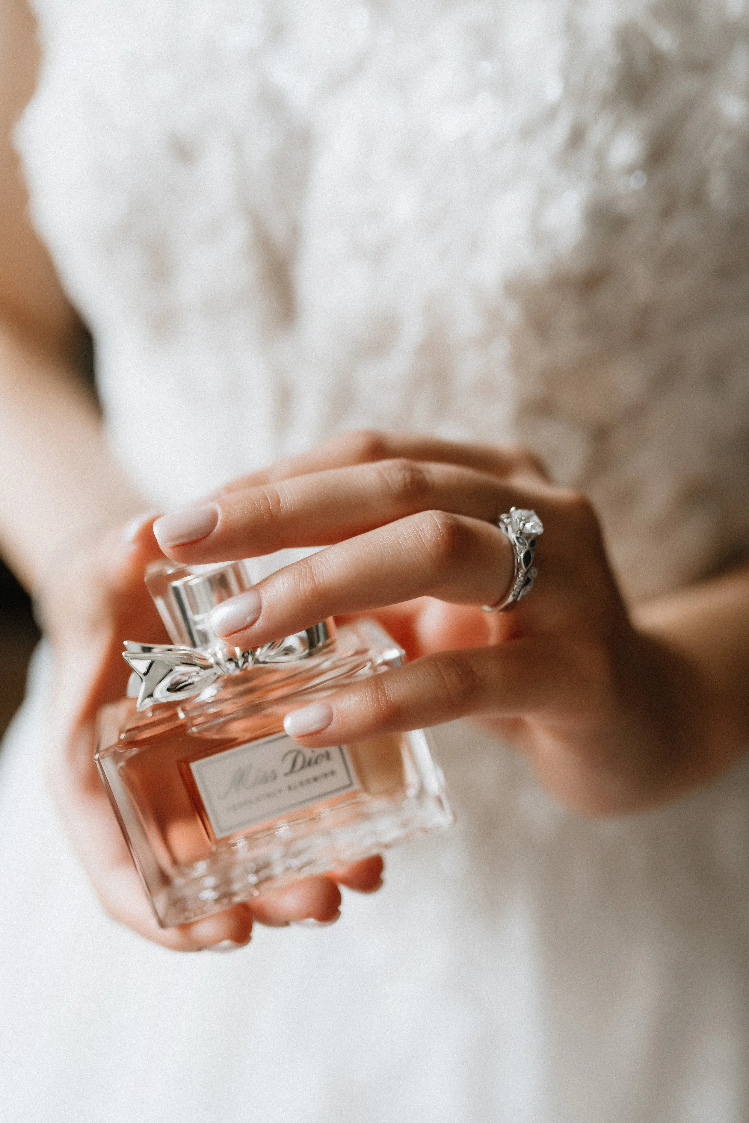 A close-up of a woman's hand holding a Miss Dior perfume bottle with a silver bow on the cap, wearing an ornate ring, against a soft, light-colored background.