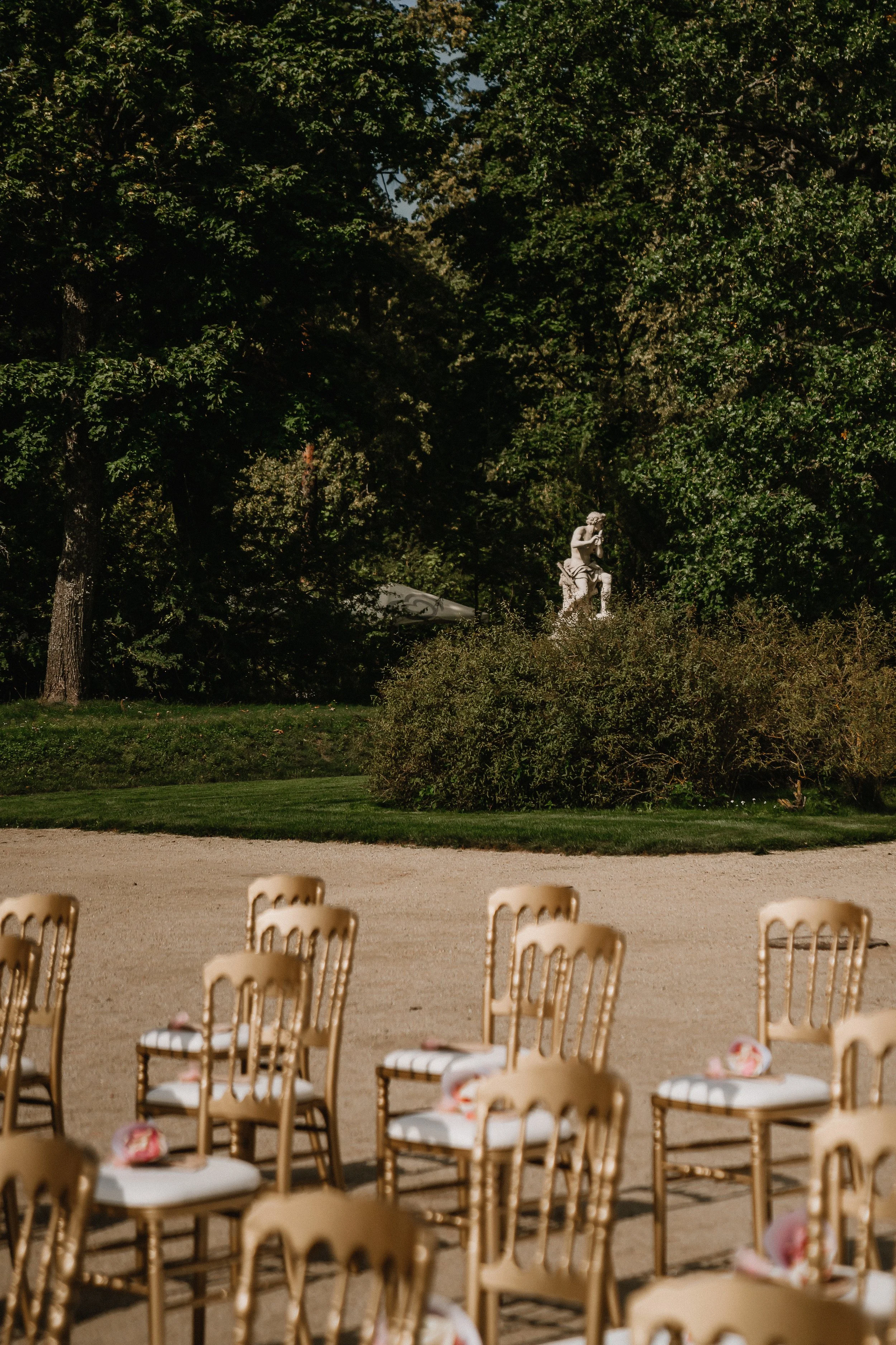 Empty chairs arranged outdoors for an event with a gravel ground, green bushes, tall trees, and a statue in the background.