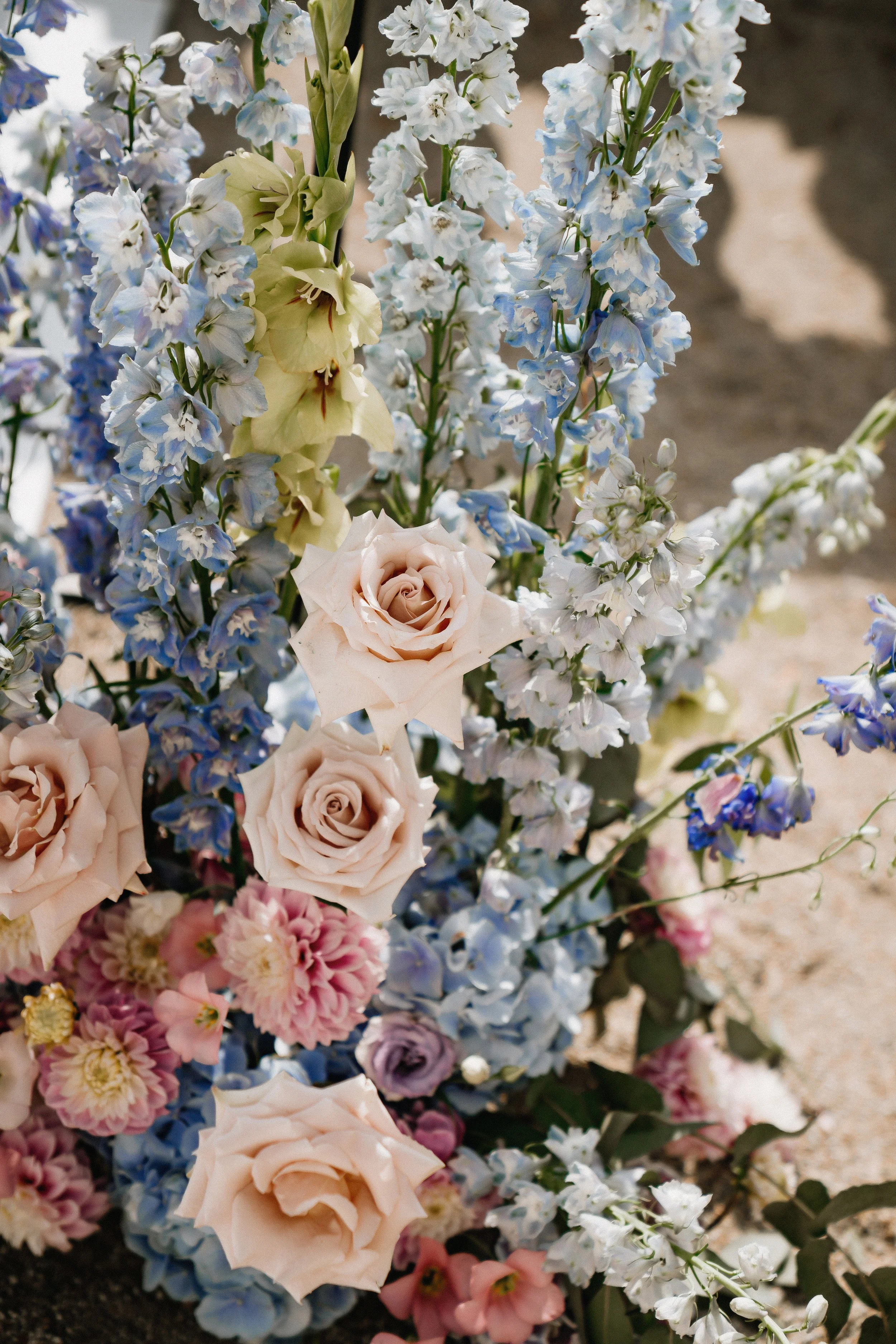 A colorful arrangement of flowers including pale pink roses, light blue delphiniums, white stock, and pink chrysanthemums, with dark soil at the base.
