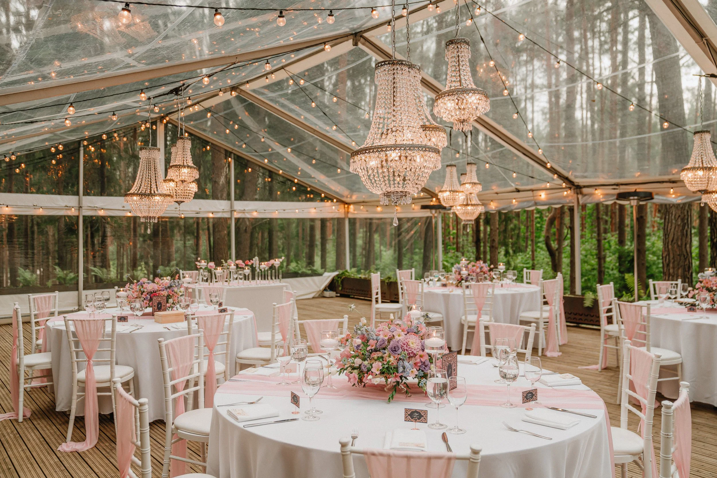 Inside a decorated outdoor tent with chandeliers, string lights, round tables with pink table runners, floral centerpieces, and white chairs, set for a celebration or wedding reception, surrounded by trees.