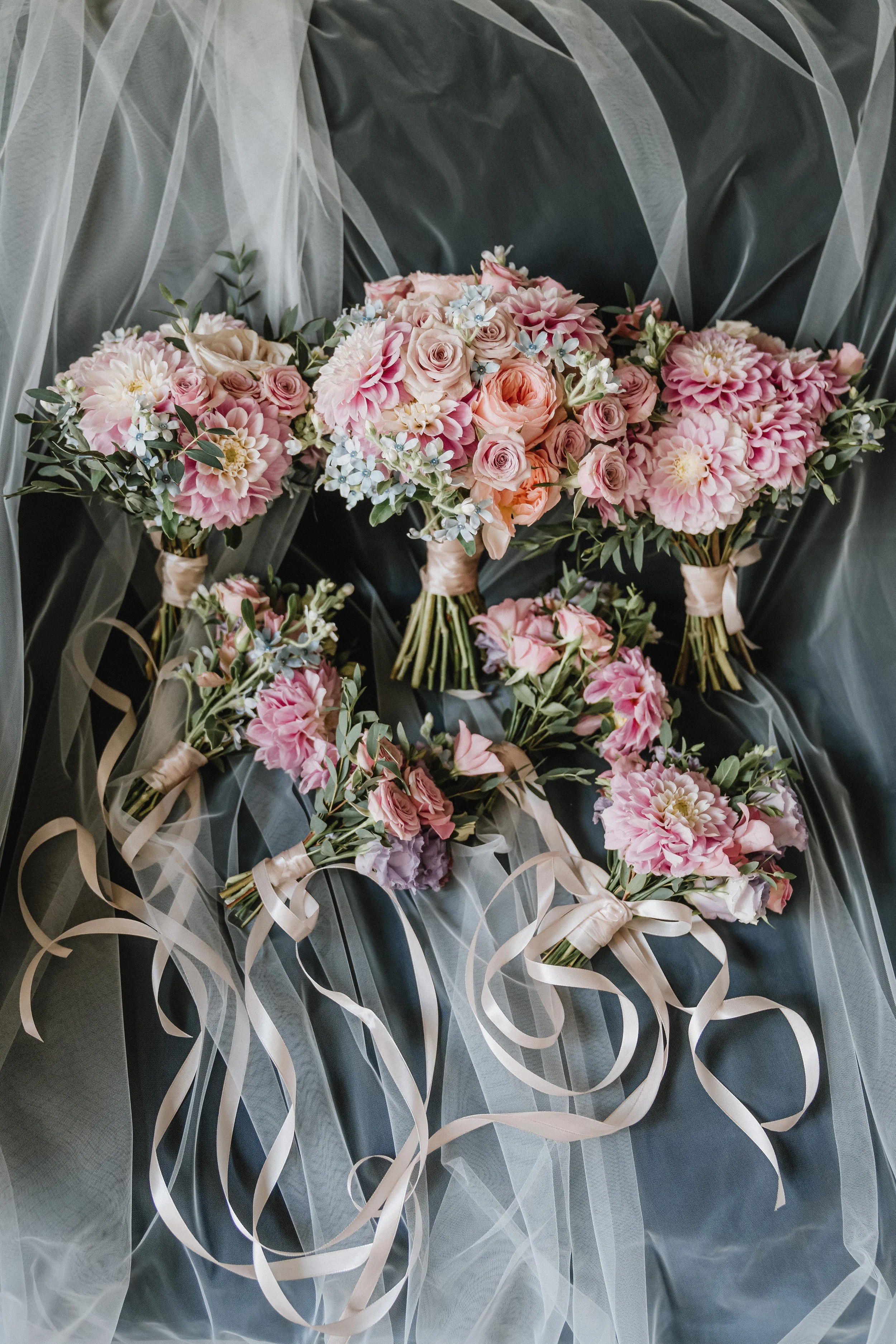 Multiple bouquets of pink and cream flowers, including roses and dahlias, with green foliage, wrapped with satin ribbons, placed against a sheer fabric backdrop.