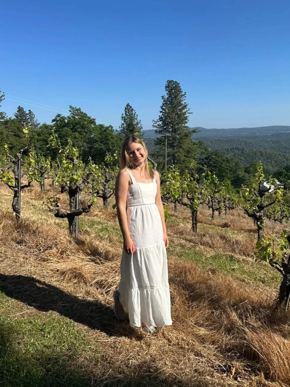A young woman standing in a vineyard on a sunny day, wearing a white dress with a backdrop of grapevines, trees, and distant hills under a clear blue sky.