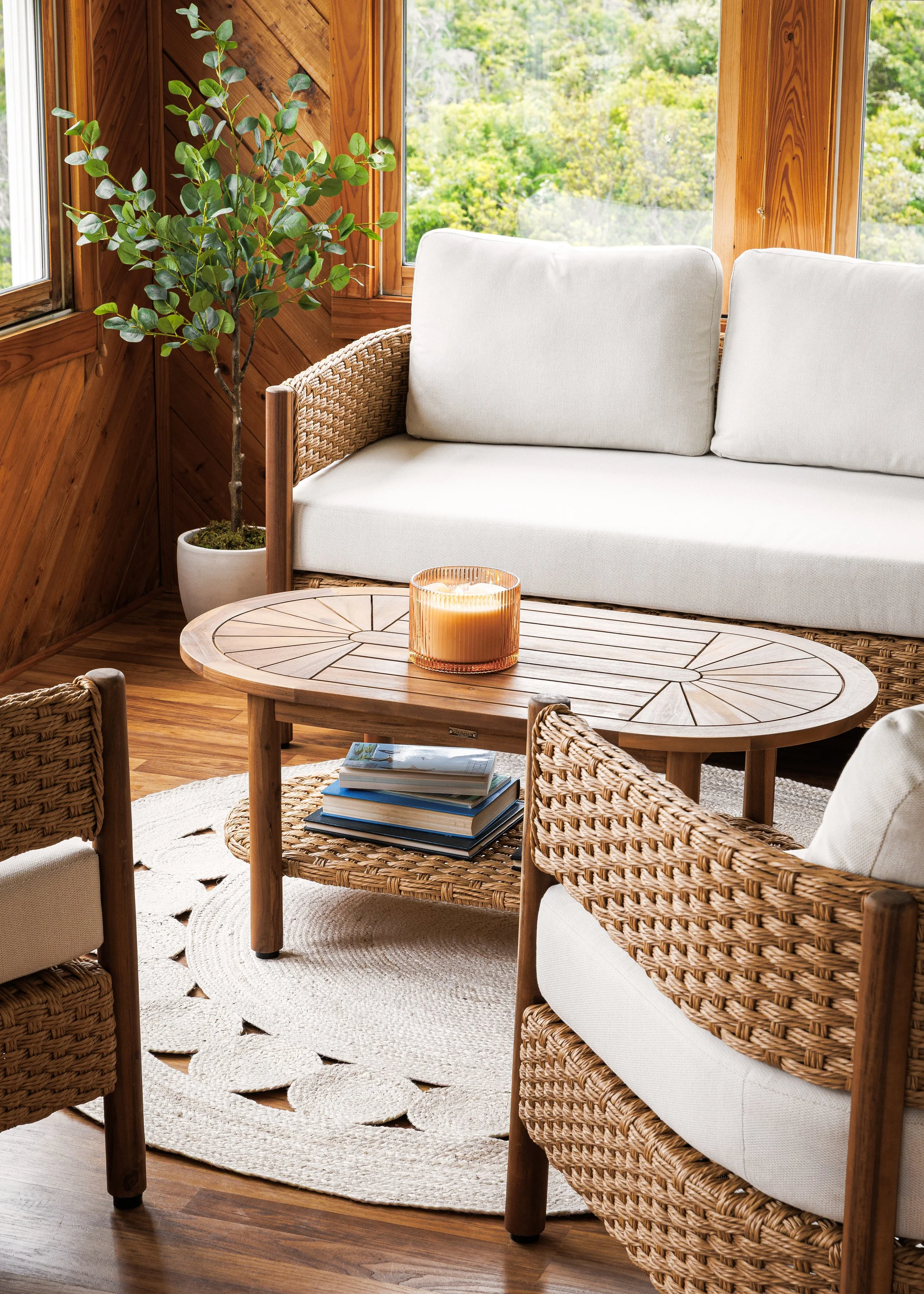 Sunroom with a white sofa, wicker armchairs, a wooden coffee table with books and a candle, a large potted plant, wood-paneled walls, and large windows showing green foliage outside.