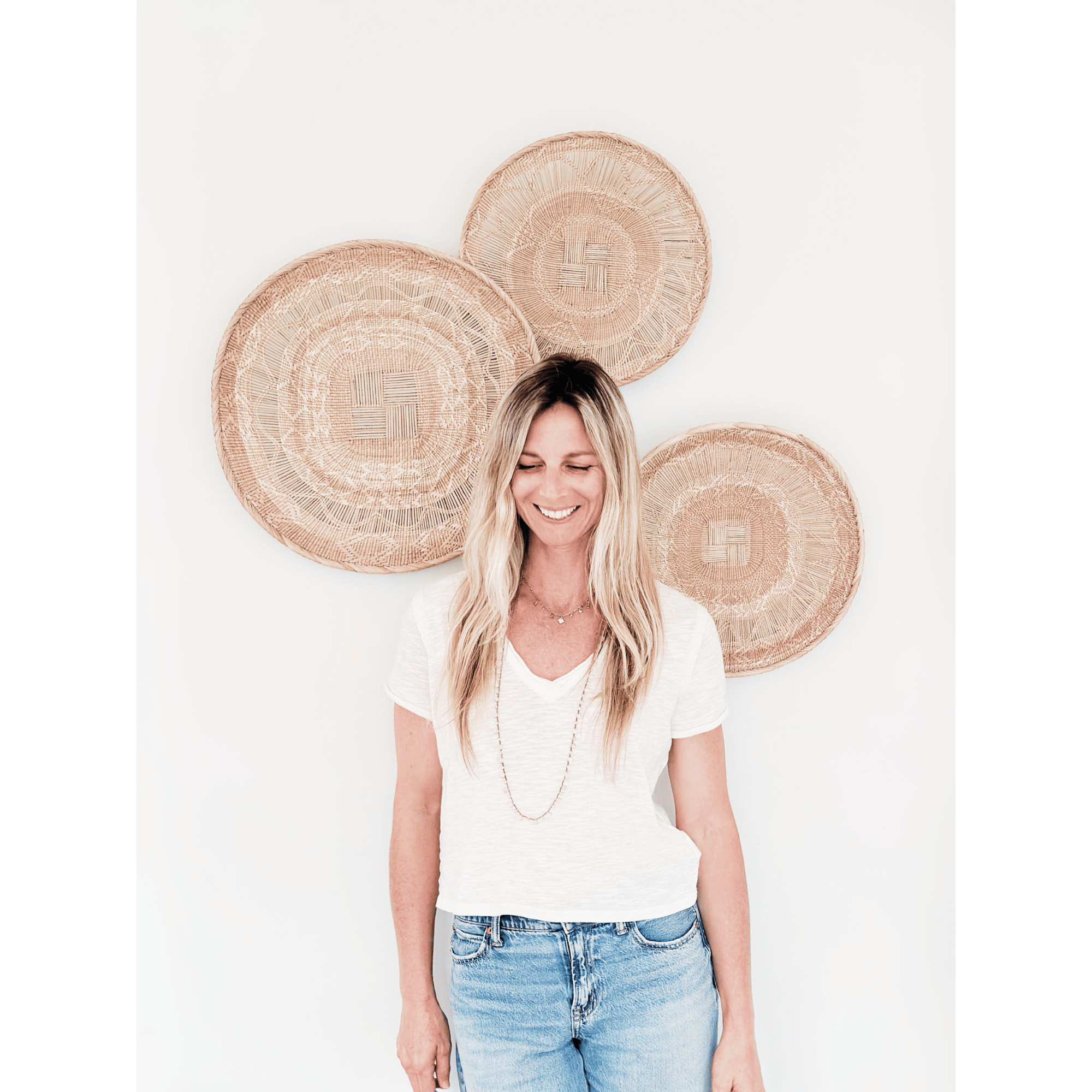 A woman with blonde hair smiling, wearing a white t-shirt and blue jeans, standing in front of a white background. Behind her are large, woven straw baskets with designs, arranged in a semi-circle.