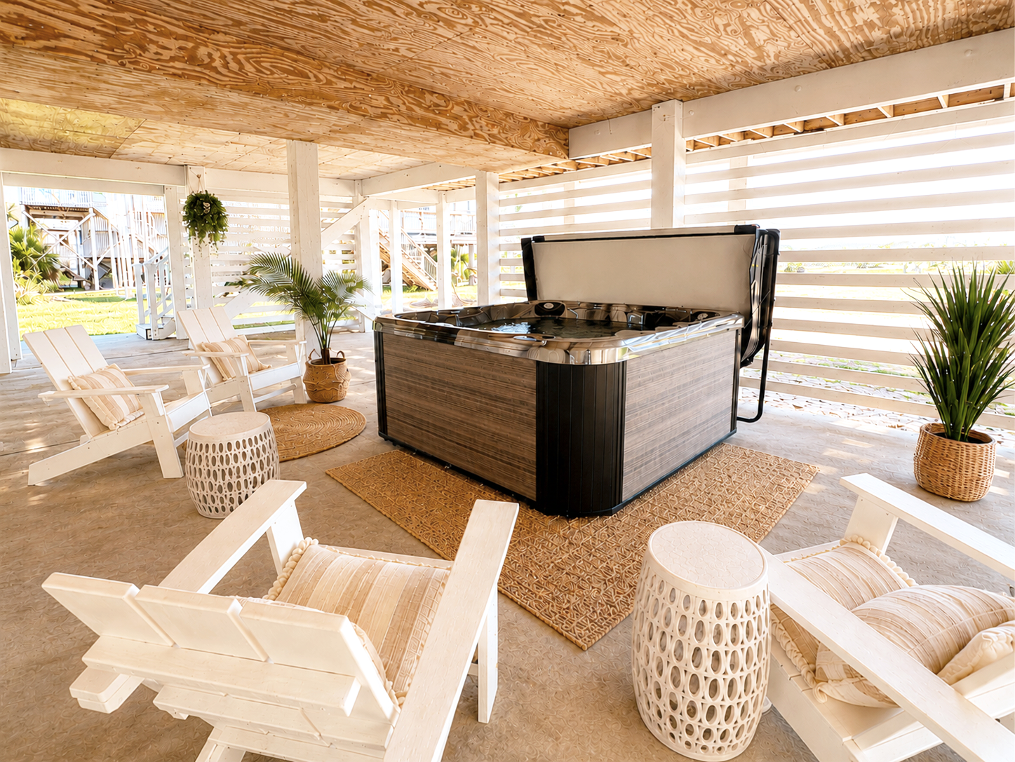 Outdoor patio with a hot tub, white wooden chairs with cushions, potted plants, and woven rugs under a wooden ceiling with white slatted walls.