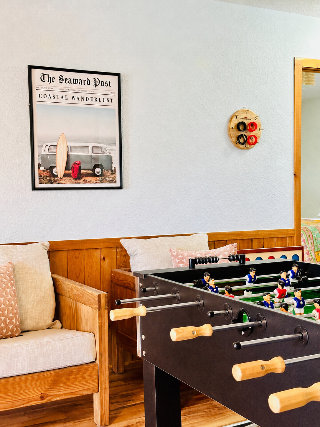 A room featuring a foosball table, a wooden bench with pillows, a framed poster of a coast-themed magazine, and a wall-mounted thermometer. The room has wooden accents and a cozy atmosphere.