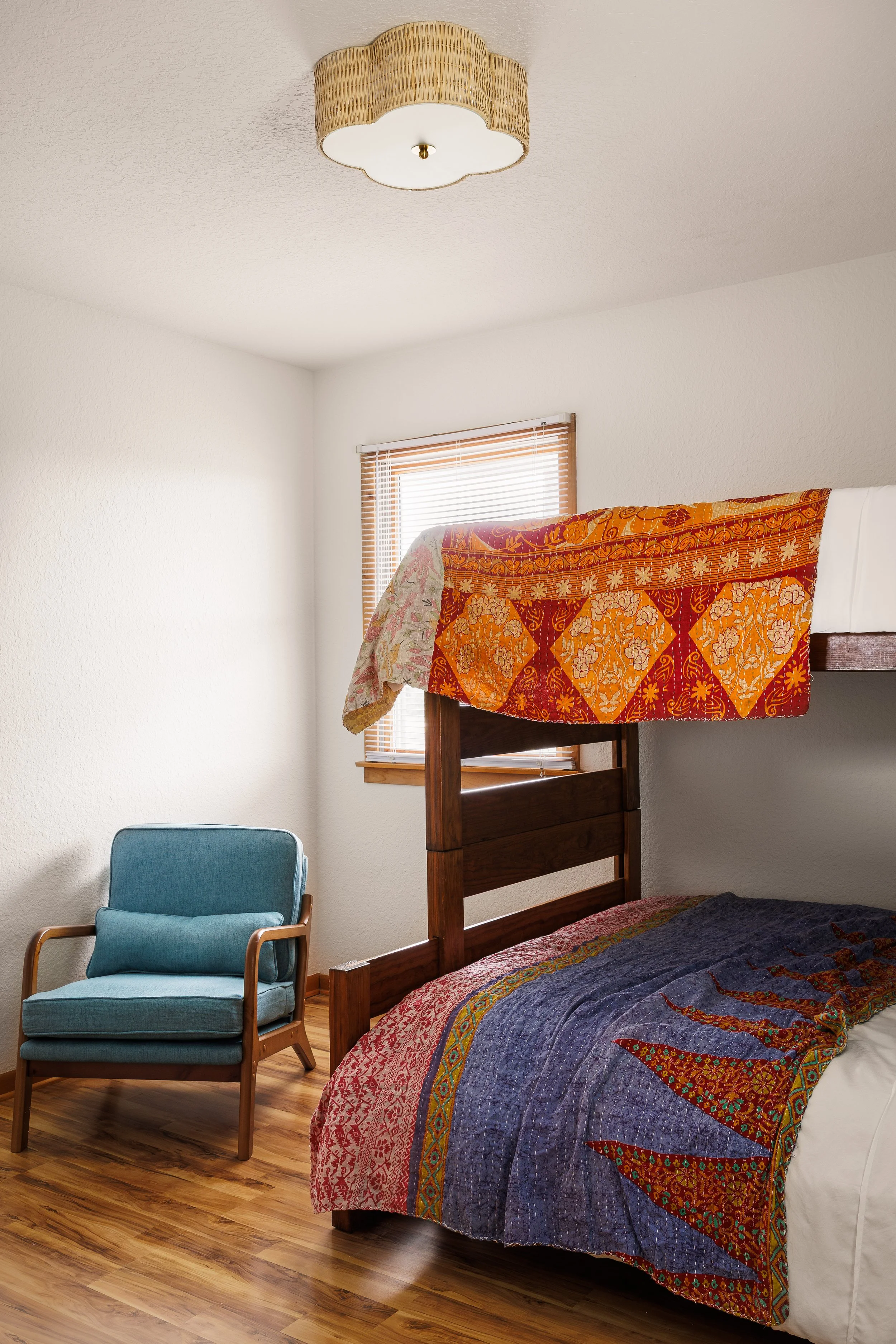 A bedroom with a wooden bunk bed covered by a colorful quilt, a blue armchair next to the bed, and a window with blinds on the wall, all on a wooden floor.