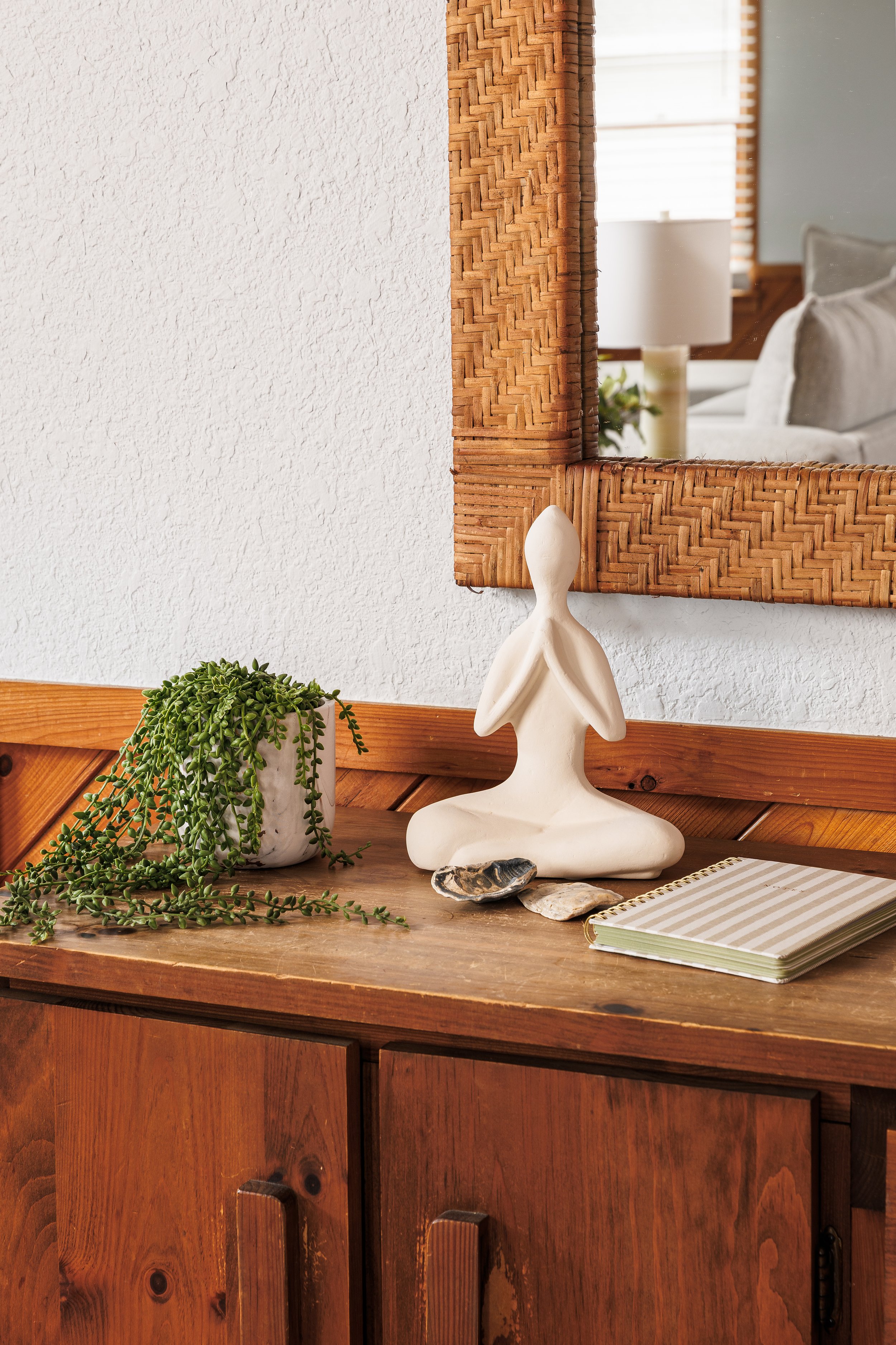 A wooden dresser with a potted trailing green plant, a white abstract sculpture, seashells, a striped notebook, and a large mirror with a woven wooden frame, reflecting a living room with a white lamp, cushions, window blinds, and a plant.
