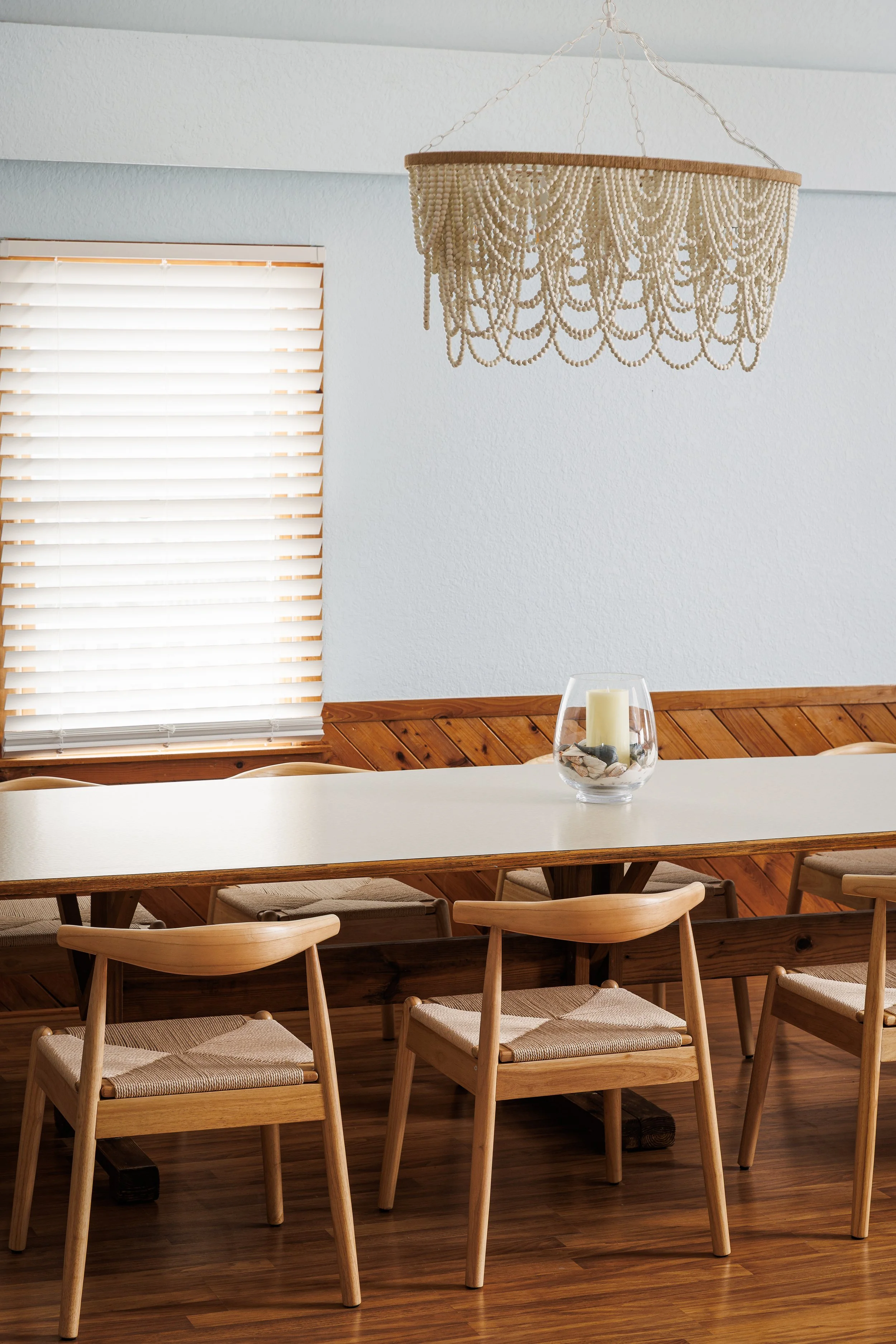 Dining room with wooden table and chairs, a glass bowl with candles and stones, a large decorative hanging light fixture, window with blinds, wood paneling, and a white wall