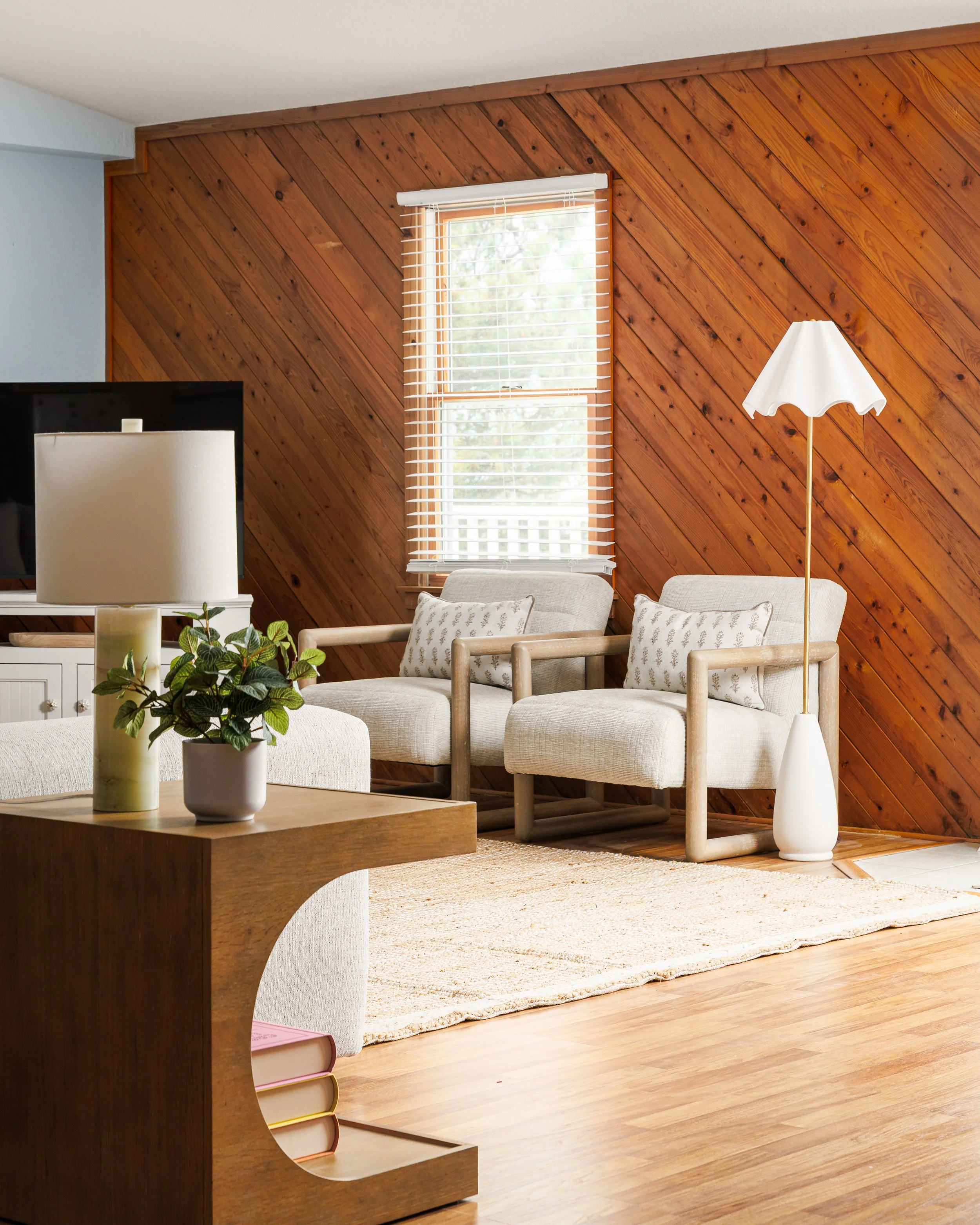 Living room with wood-paneled wall, white window blinds, beige upholstered furniture, a white floor lamp, and a wooden coffee table with potted plants.
