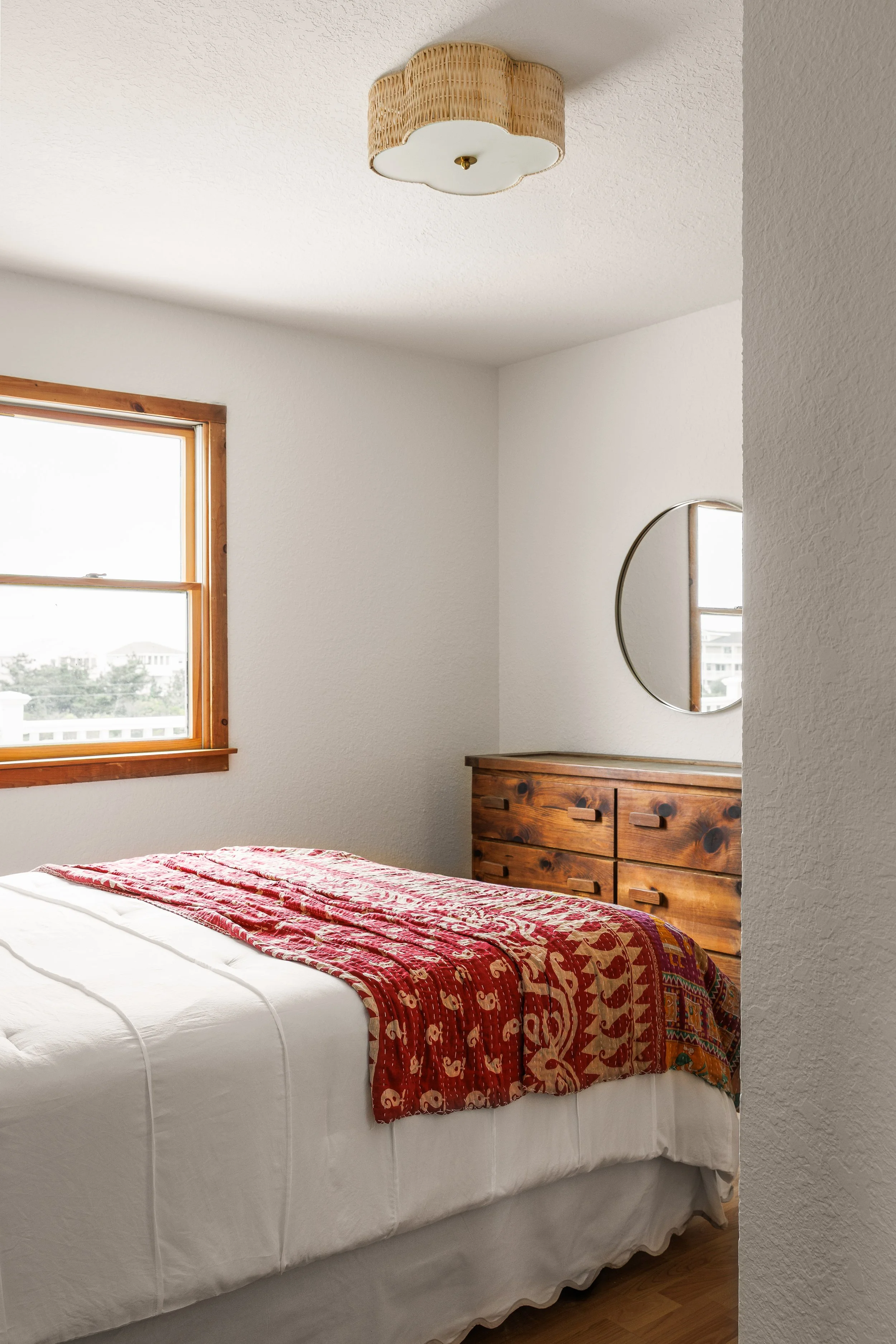 A bedroom with a bed covered in white linens and a red and gold patterned quilt. There is a wooden dresser with a mirror on top, and a window with wooden trim letting in natural light.