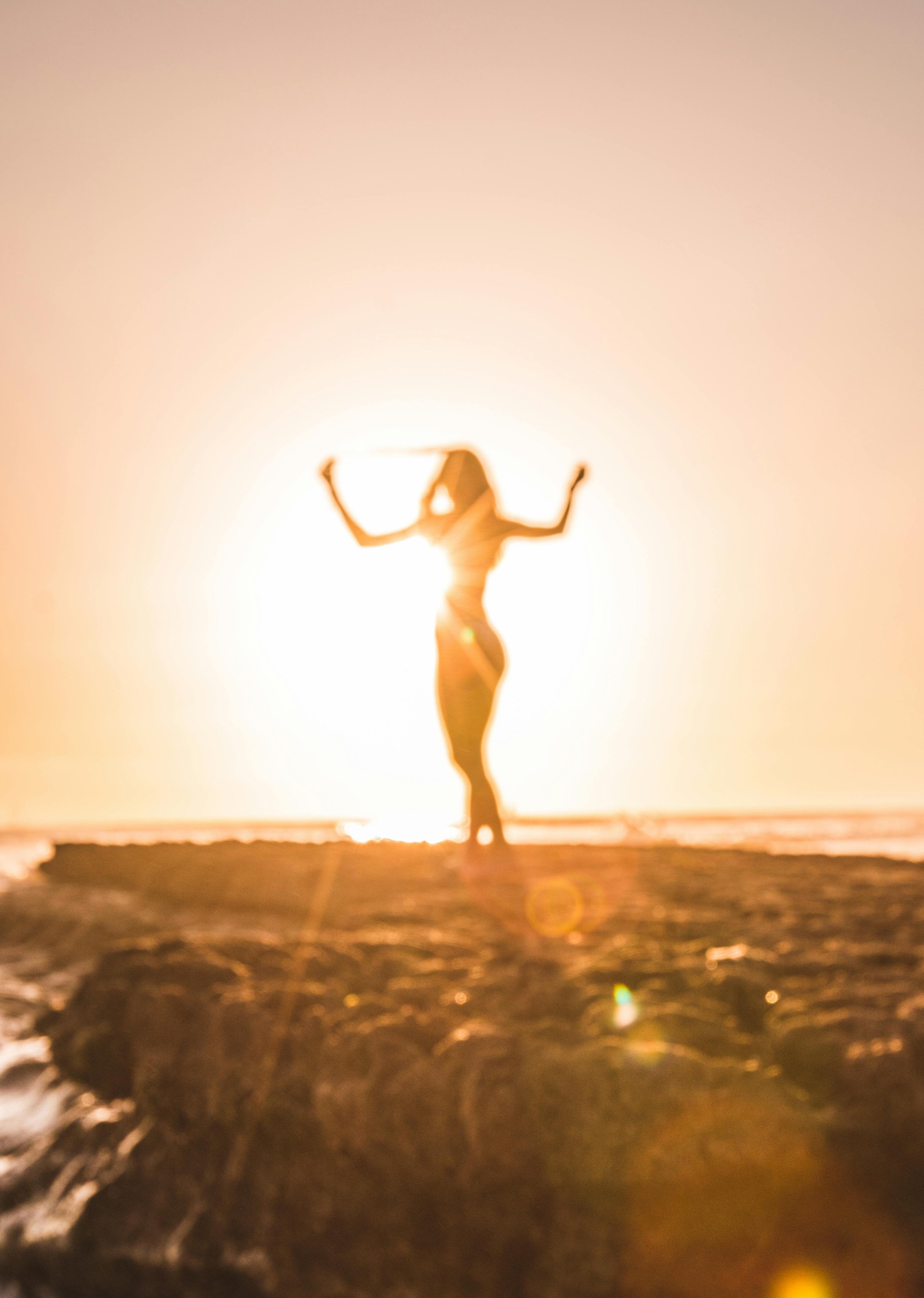 A person practicing yoga or tai chi outdoors at sunset, standing on a beach or shoreline with water and rocks, silhouette against the bright sun.