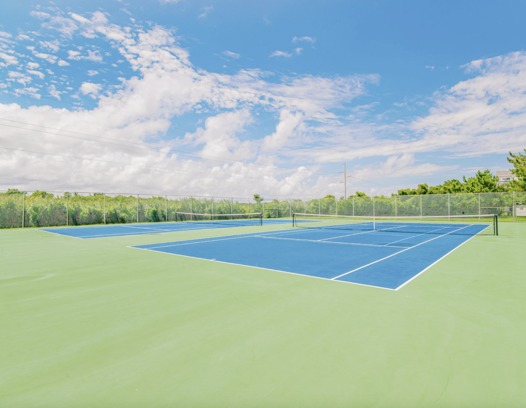 An empty outdoor tennis court with blue playing surfaces, surrounded by a chain-link fence, under a partly cloudy sky with green trees in the background.