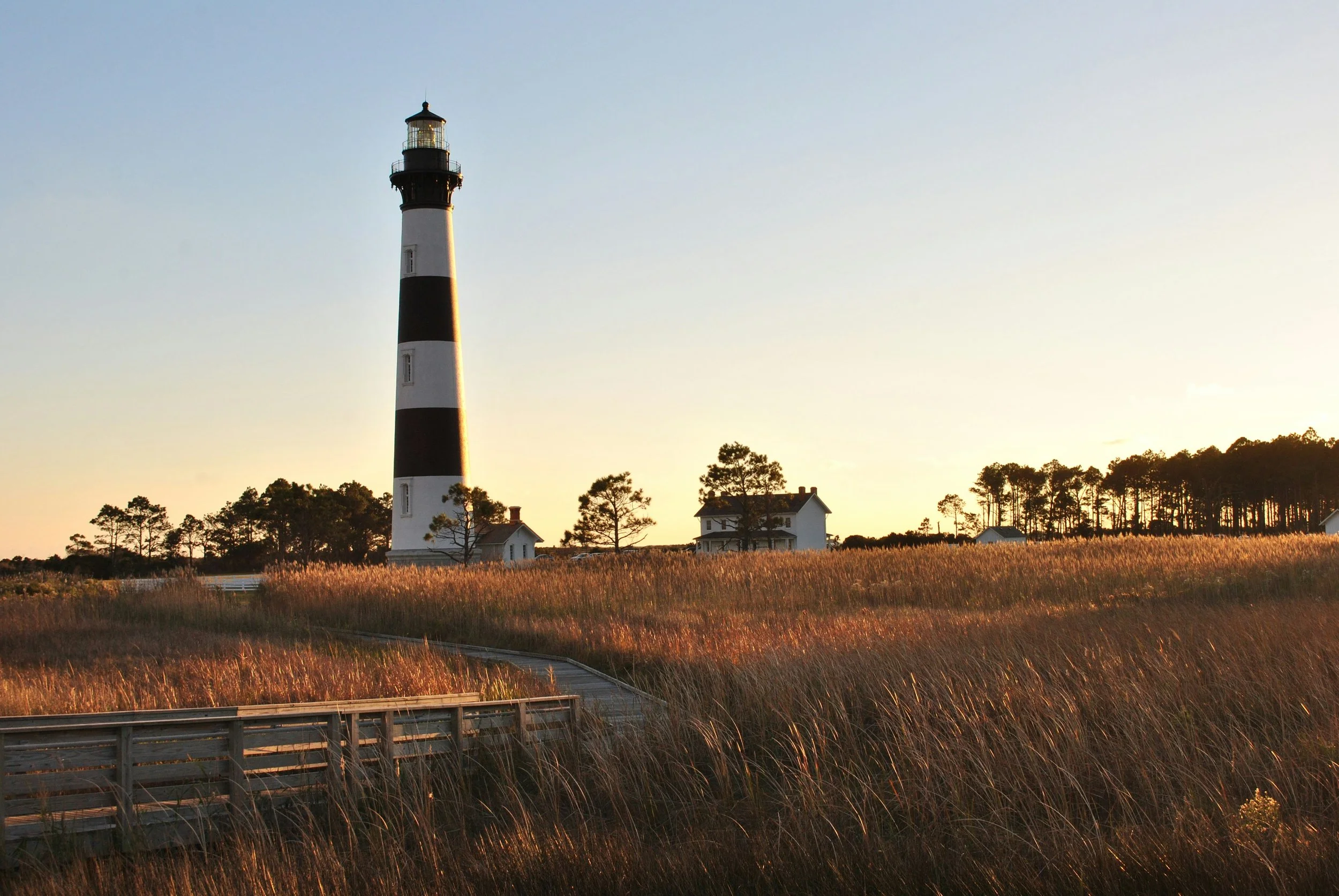 A lighthouse with black and white horizontal stripes stands among trees and houses in a grassy field during sunset.