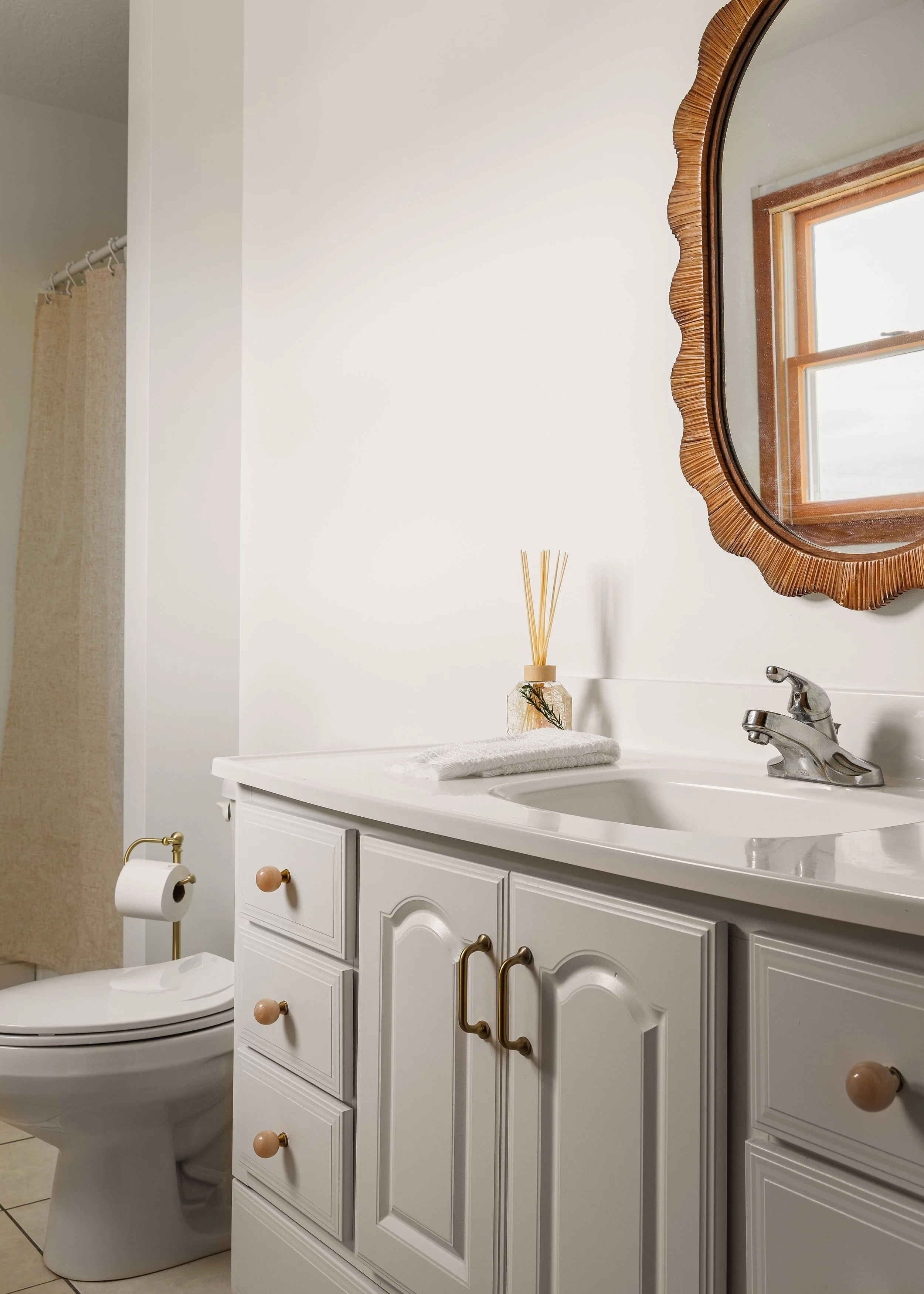 A bathroom with a white vanity and cabinet, a framed mirror, a potted plant fragrance diffuser, a toilet paper holder with a roll, and a window with wooden trim.