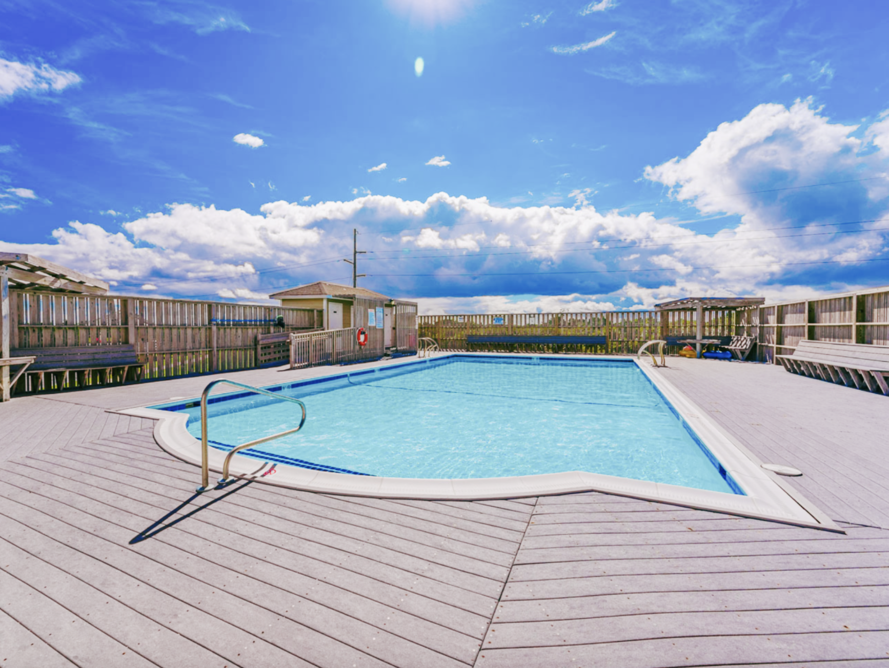 Public outdoor swimming pool with wooden deck, benches, fencing, and a small building, under a partly cloudy blue sky.