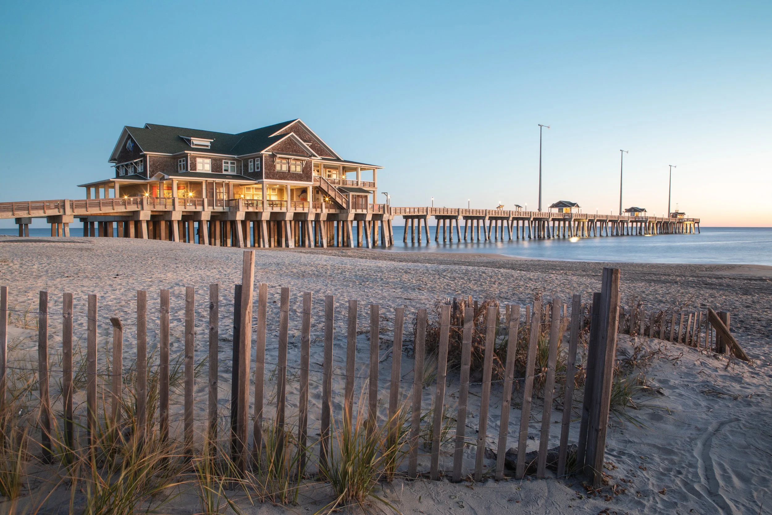 A wooden pier extends over the water at sunset, with a house at the end and smaller structures along the pier, fenced sandy beach in the foreground, and clear blue sky.