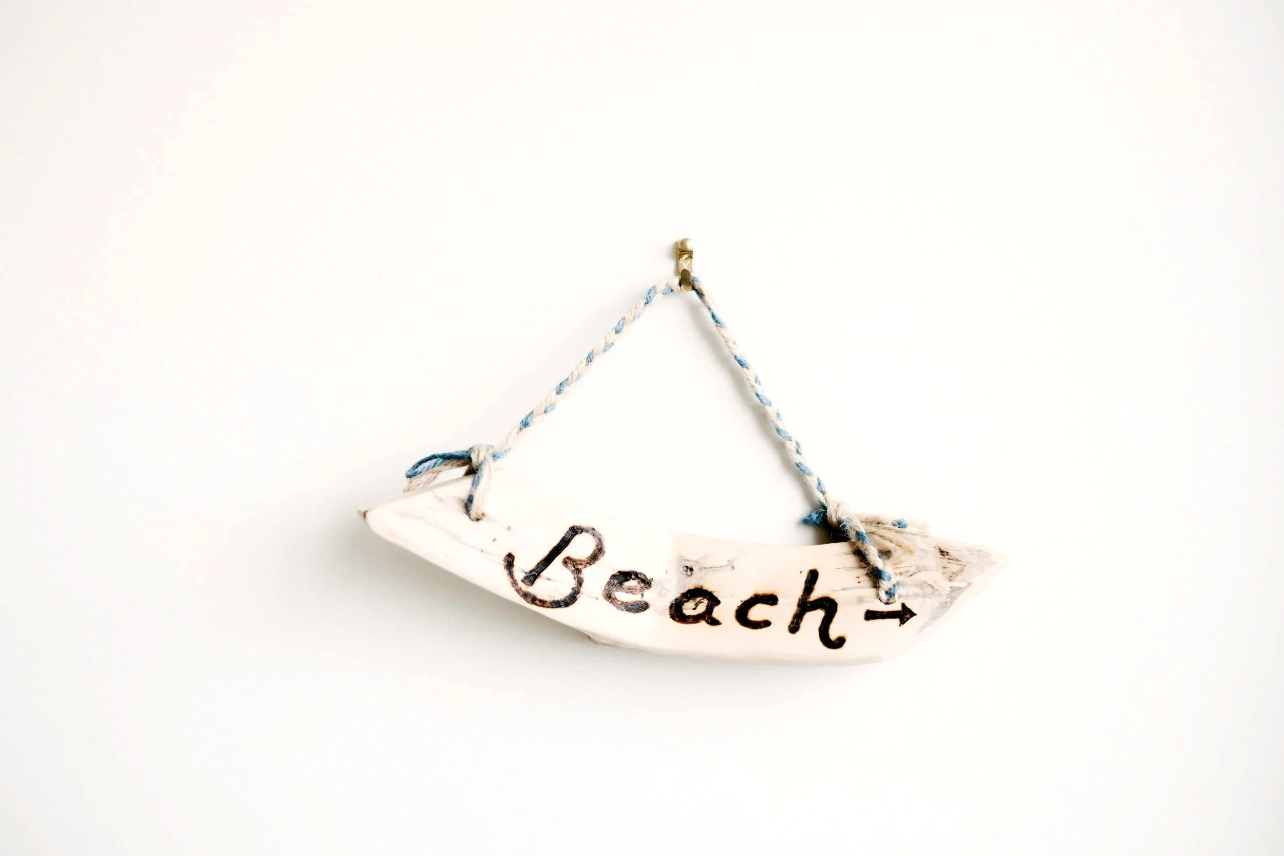 Wooden boat-shaped sign with the word 'Beach' painted on it, hanging from a blue and white twisted rope against a plain white background.