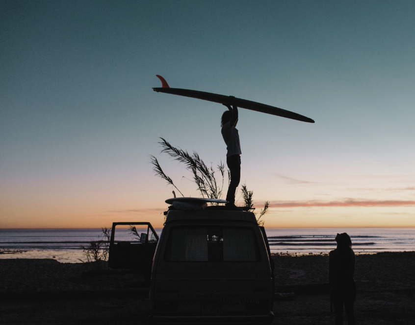 Silhouette of a person standing on top of a vehicle holding a surfboard, with a beach and sunset in the background.