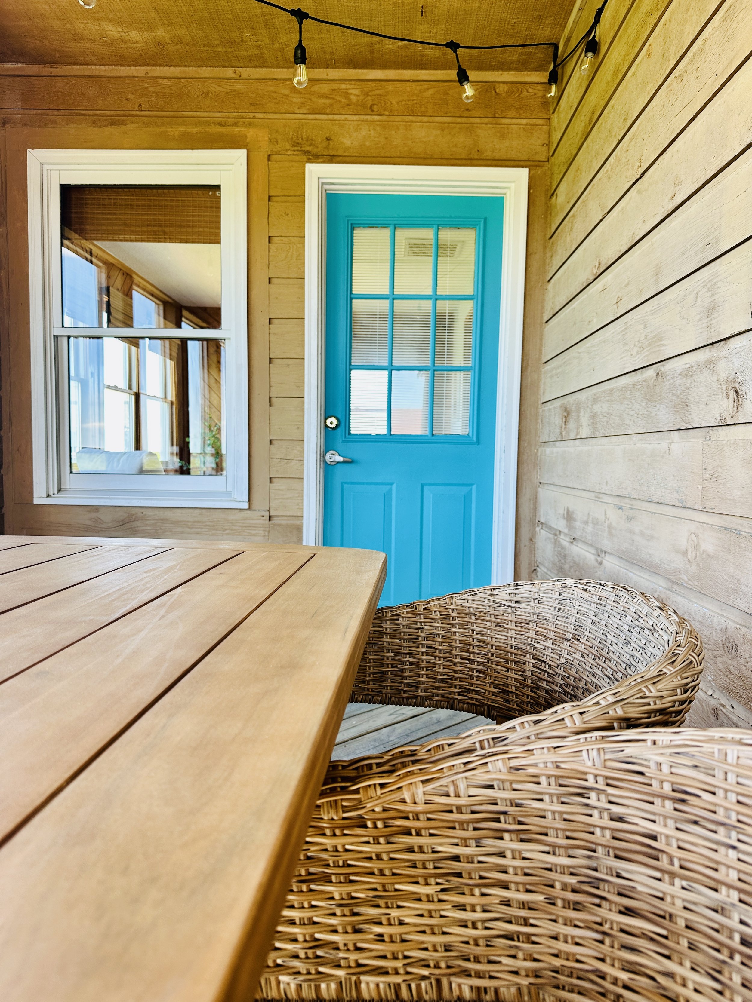 Wooden porch with a blue door, window, wicker chair, and string lights hanging from the ceiling.
