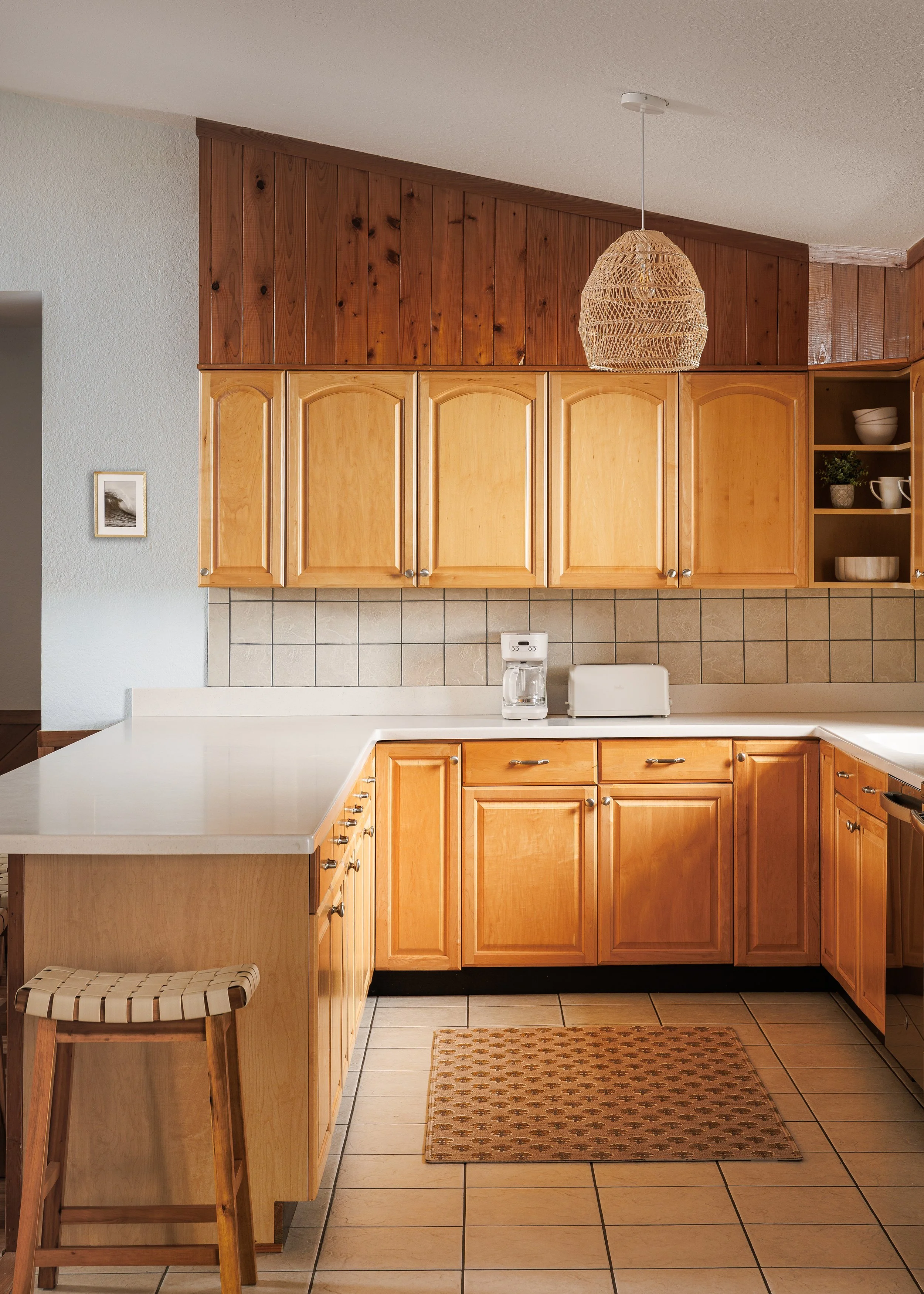 A kitchen with wooden cabinets, beige tiled backsplash, a white countertop, a coffee maker, a bread box, open shelves with bowls and mugs, a wooden stool, a patterned rug, and a pendant light made of woven material hanging from the ceiling.
