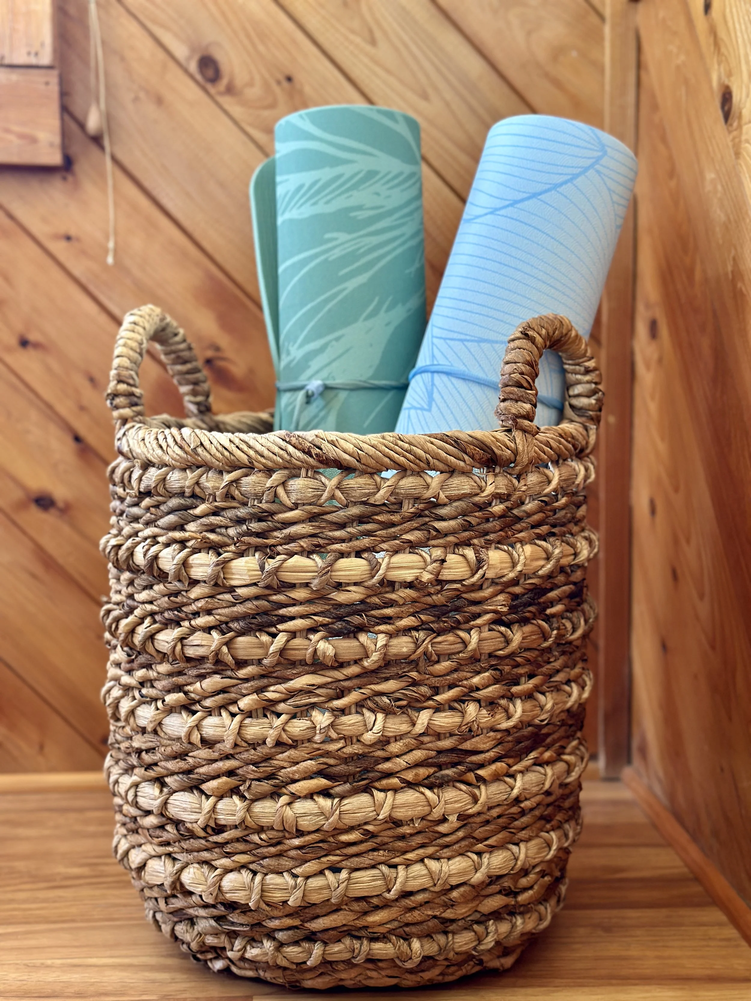 A woven wicker basket holding two rolled-up yoga mats, one green with leaf patterns and one blue with leaf patterns, sitting on a wooden floor with a wooden wall in the background.