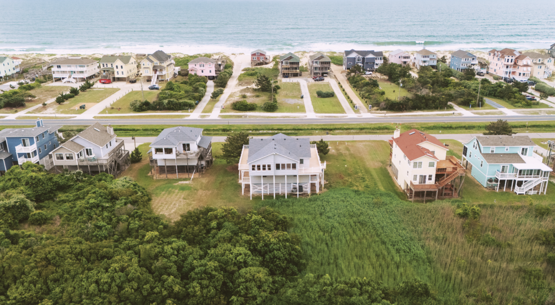 Aerial view of houses along a shoreline, with the ocean in the background, grassy areas, trees, and a road running through the scene.