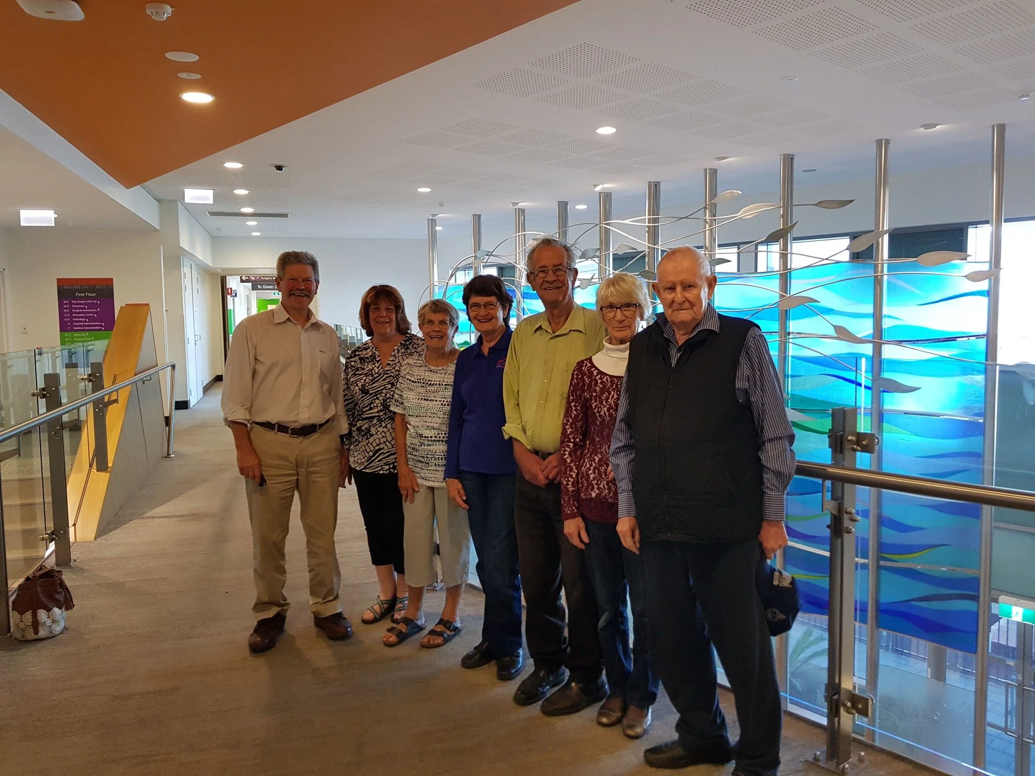 Seven elderly people standing together in a hospital or medical center hallway, smiling for a group photo with a colorful wave mural behind them.
