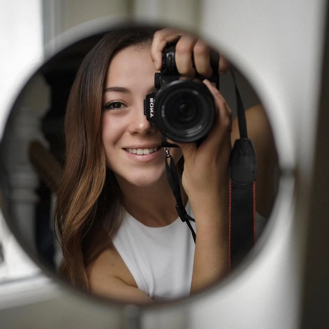 A young woman with long brown hair smiling while taking a selfie in a mirror with a Canon camera, seen through a round mirror frame.